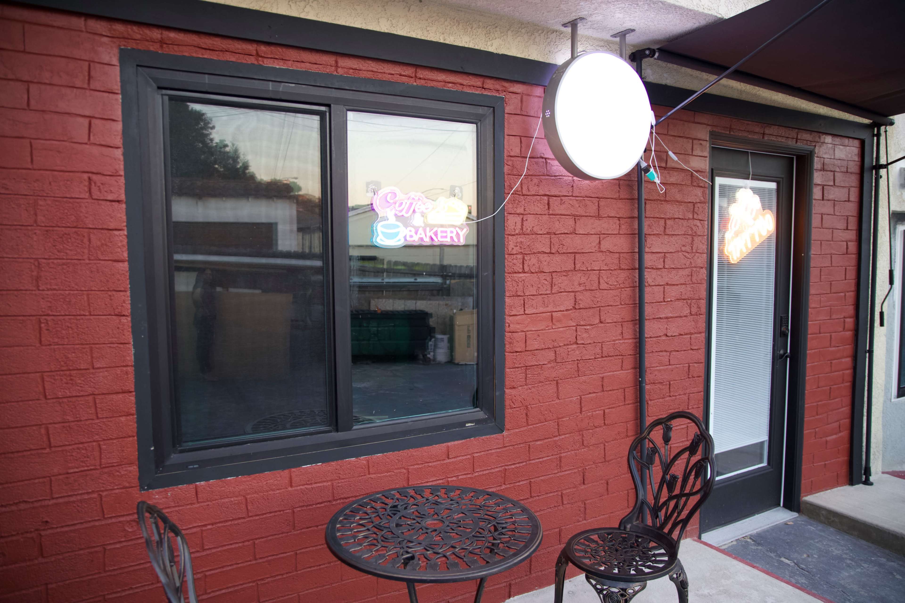 A red brick wall features a window with a neon "Bakery" sign and a small outdoor table with chairs.