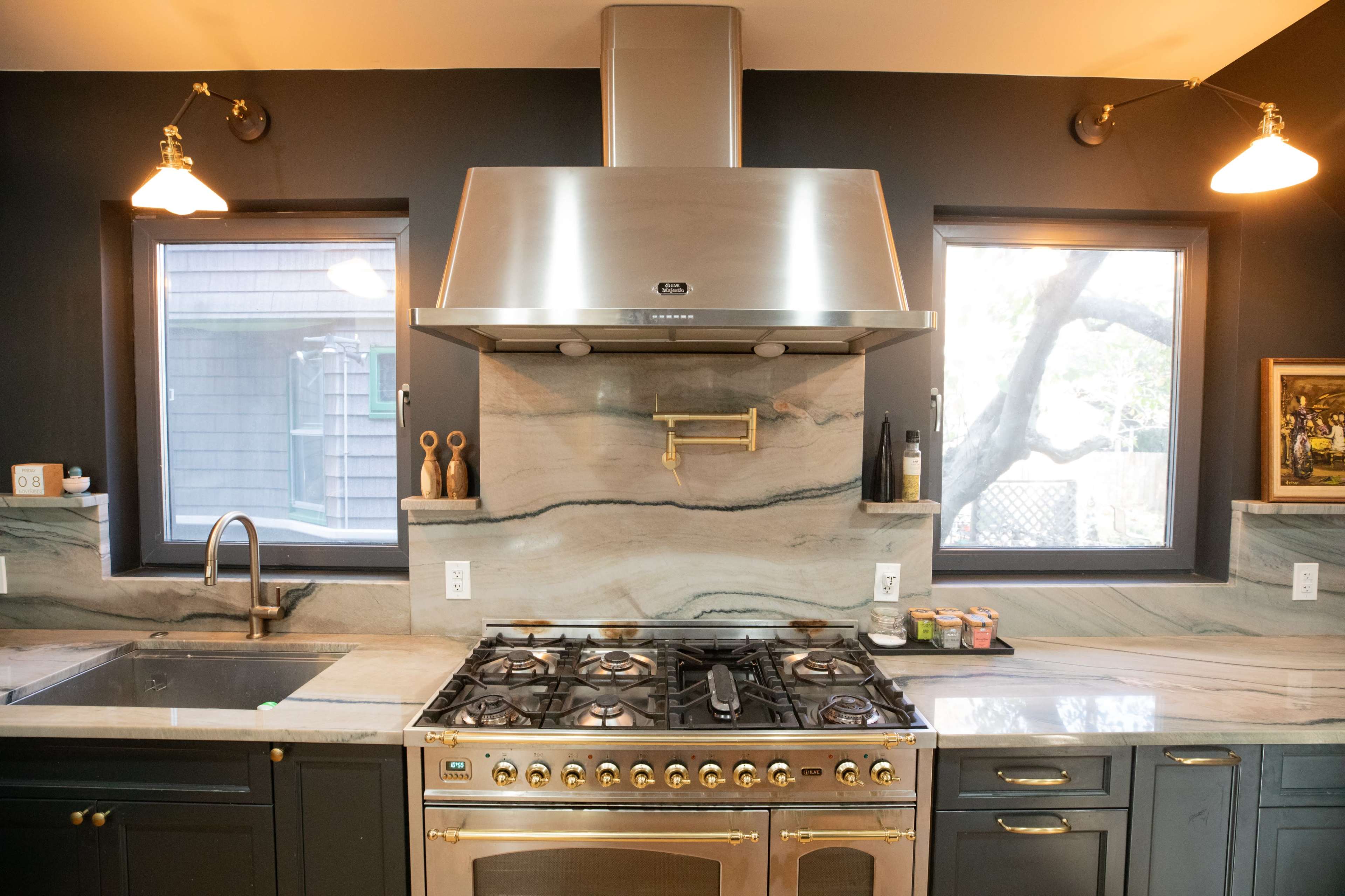 A modern kitchen features a large gas stove with a metallic hood, marble countertops, and two windows overlooking a tree.