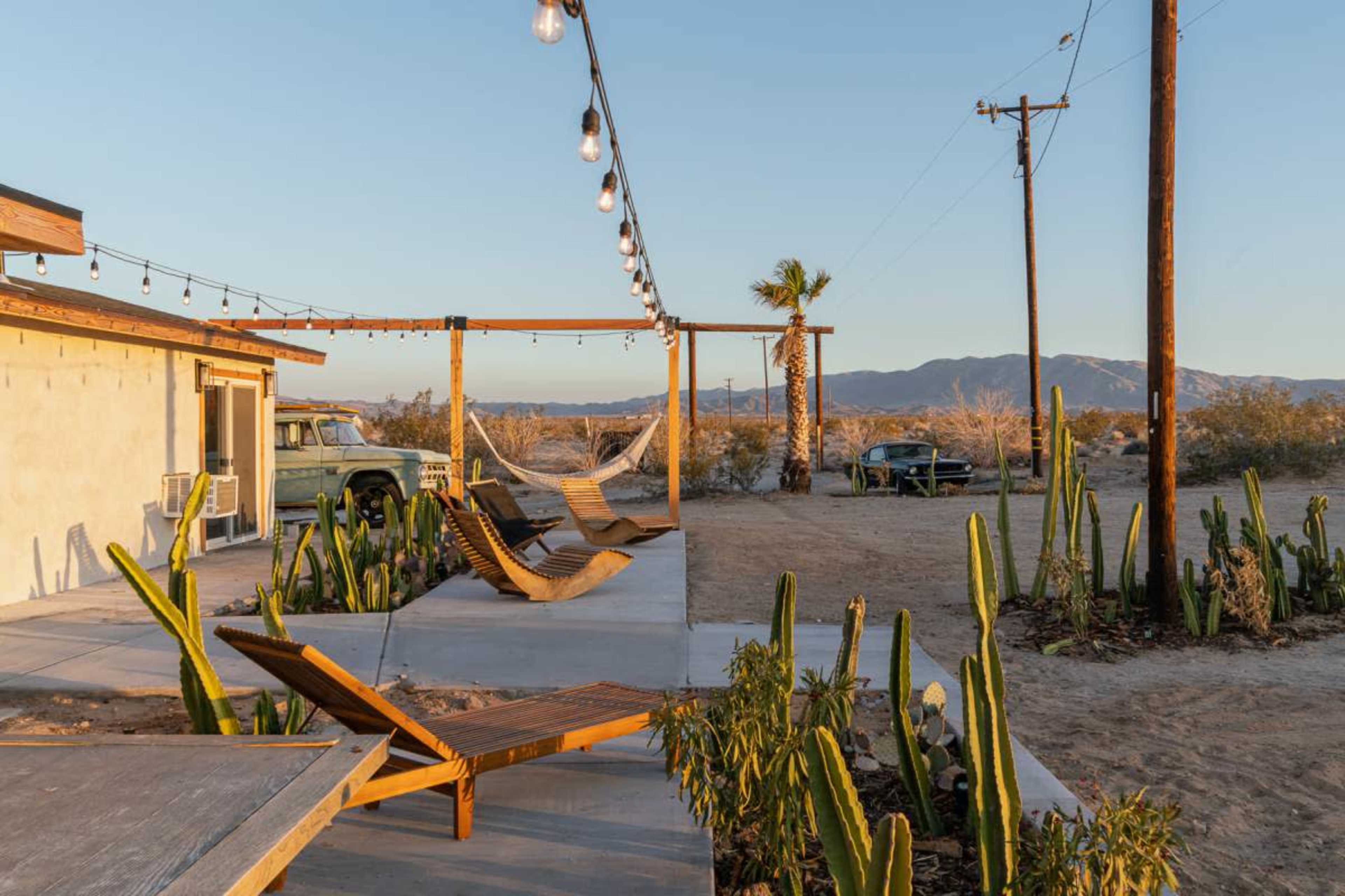 The image shows a desert landscape with a modern building, wooden lounge chairs, a hammock, and cacti, under a string of lights, with mountains in the background.