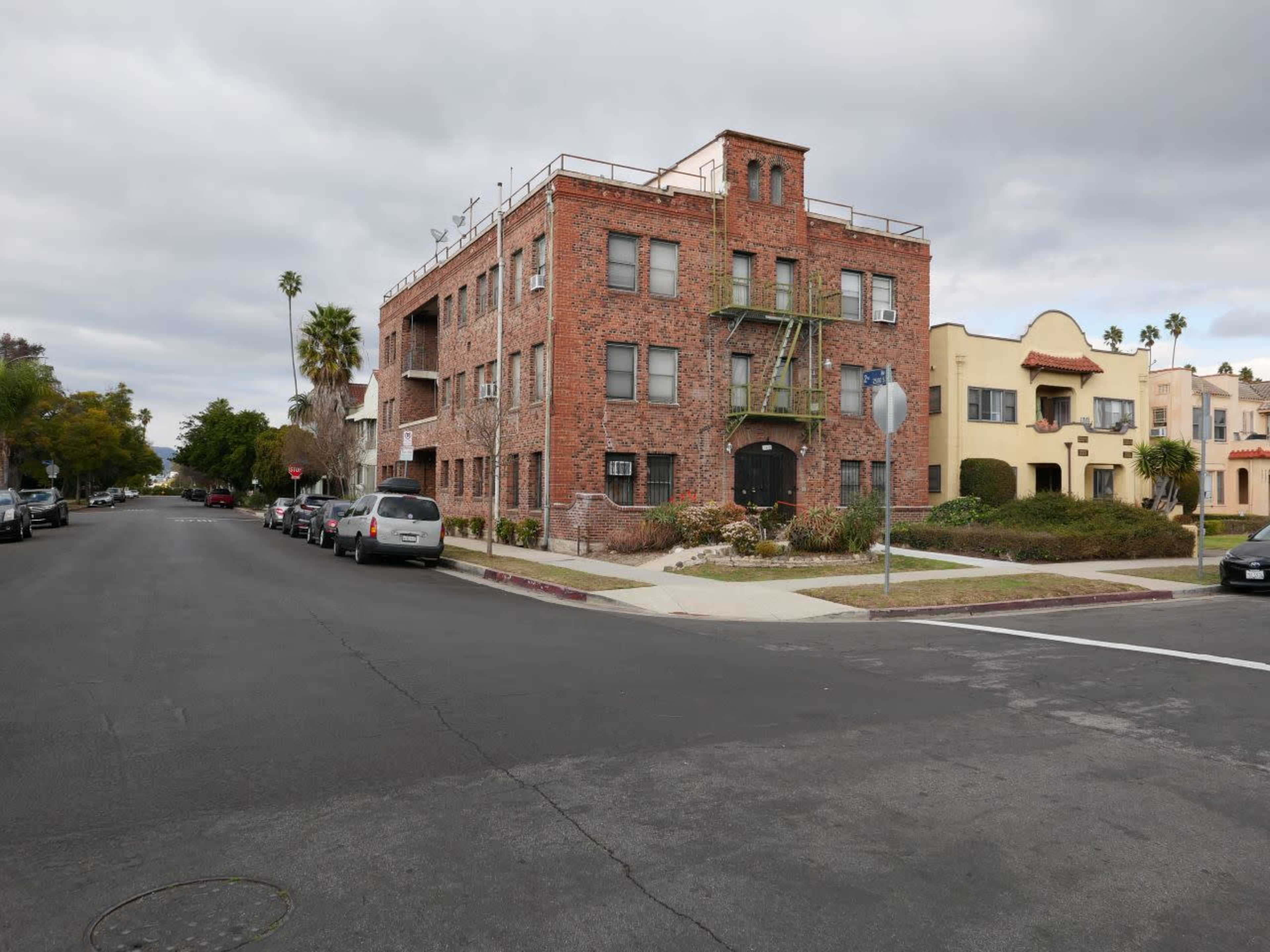 The image shows a brick apartment building located at the intersection of a residential street, with parked cars lined along both sides of the road.