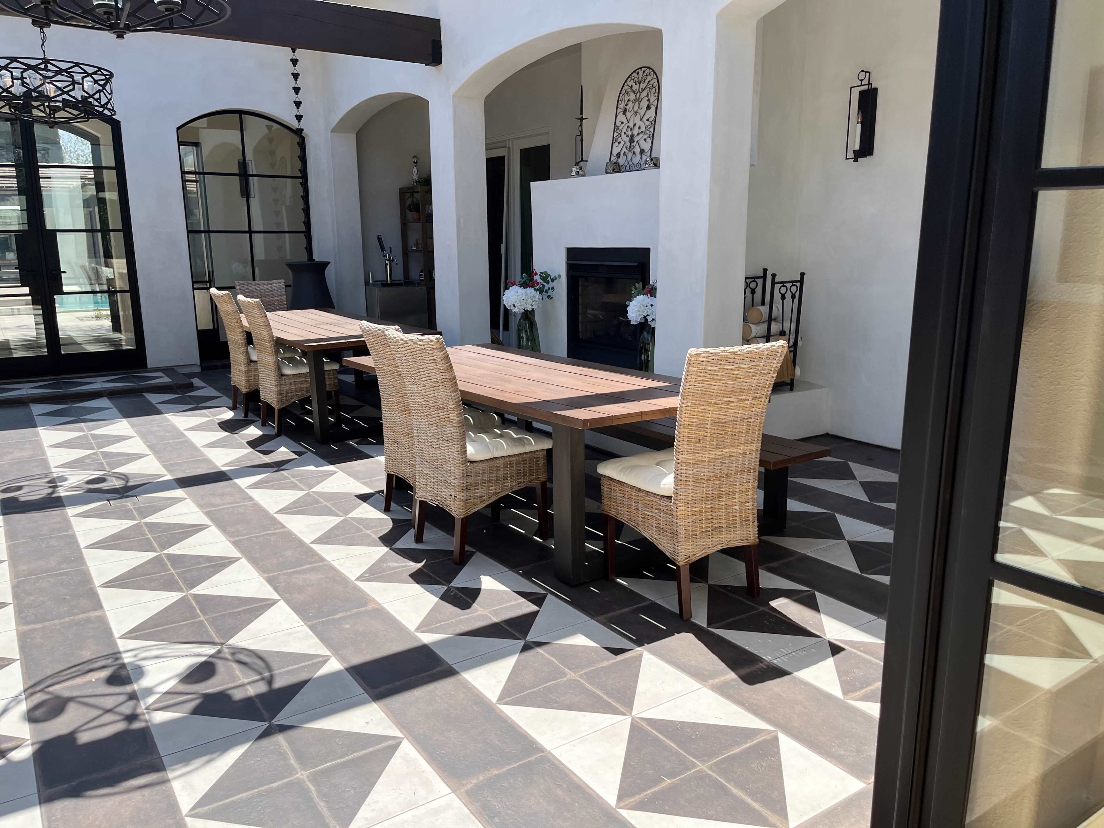 A spacious outdoor dining area features a long wooden table with wicker chairs, set on a patterned tiled floor.