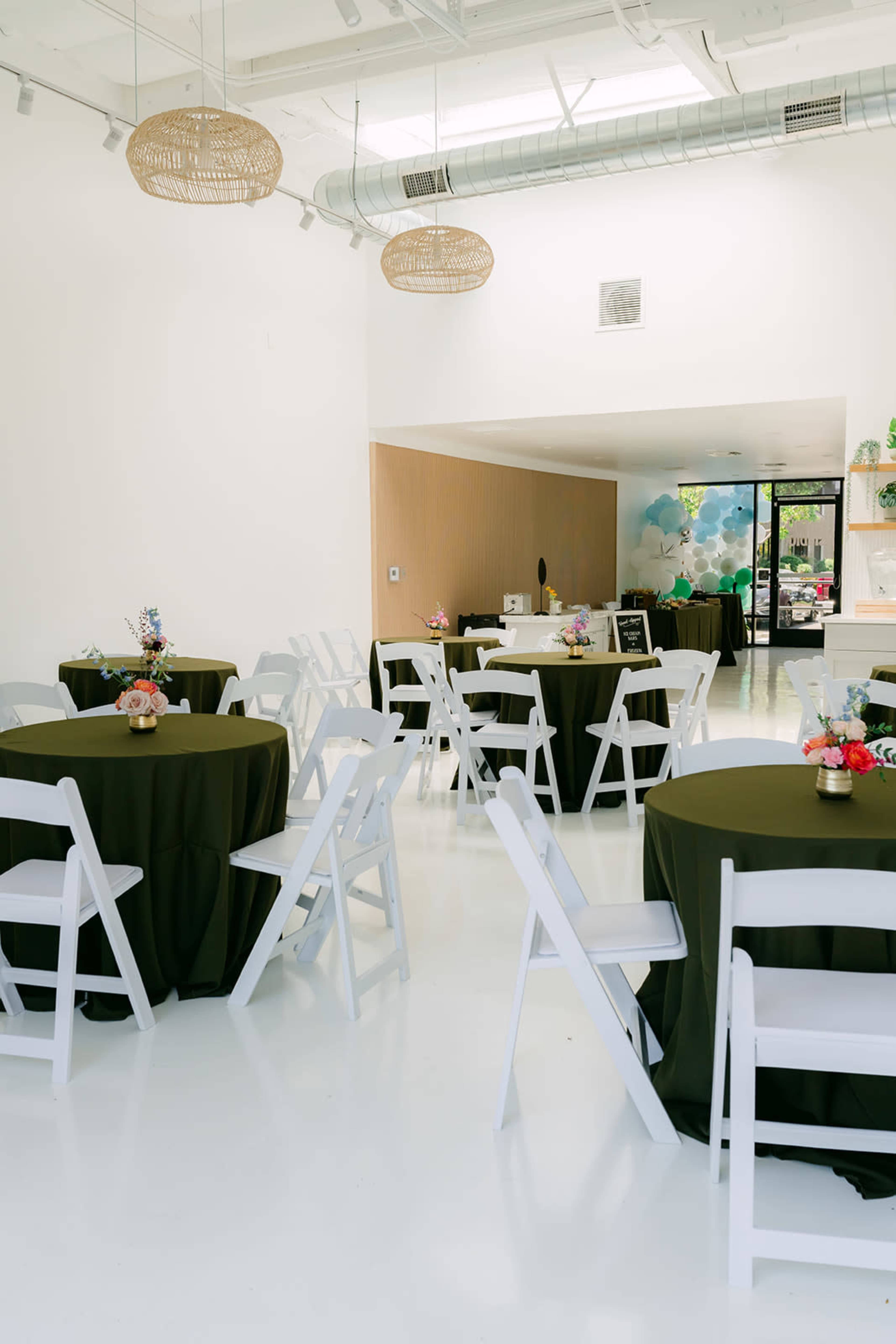 The image shows a well-lit event space with round tables covered in dark green tablecloths and surrounded by white folding chairs.