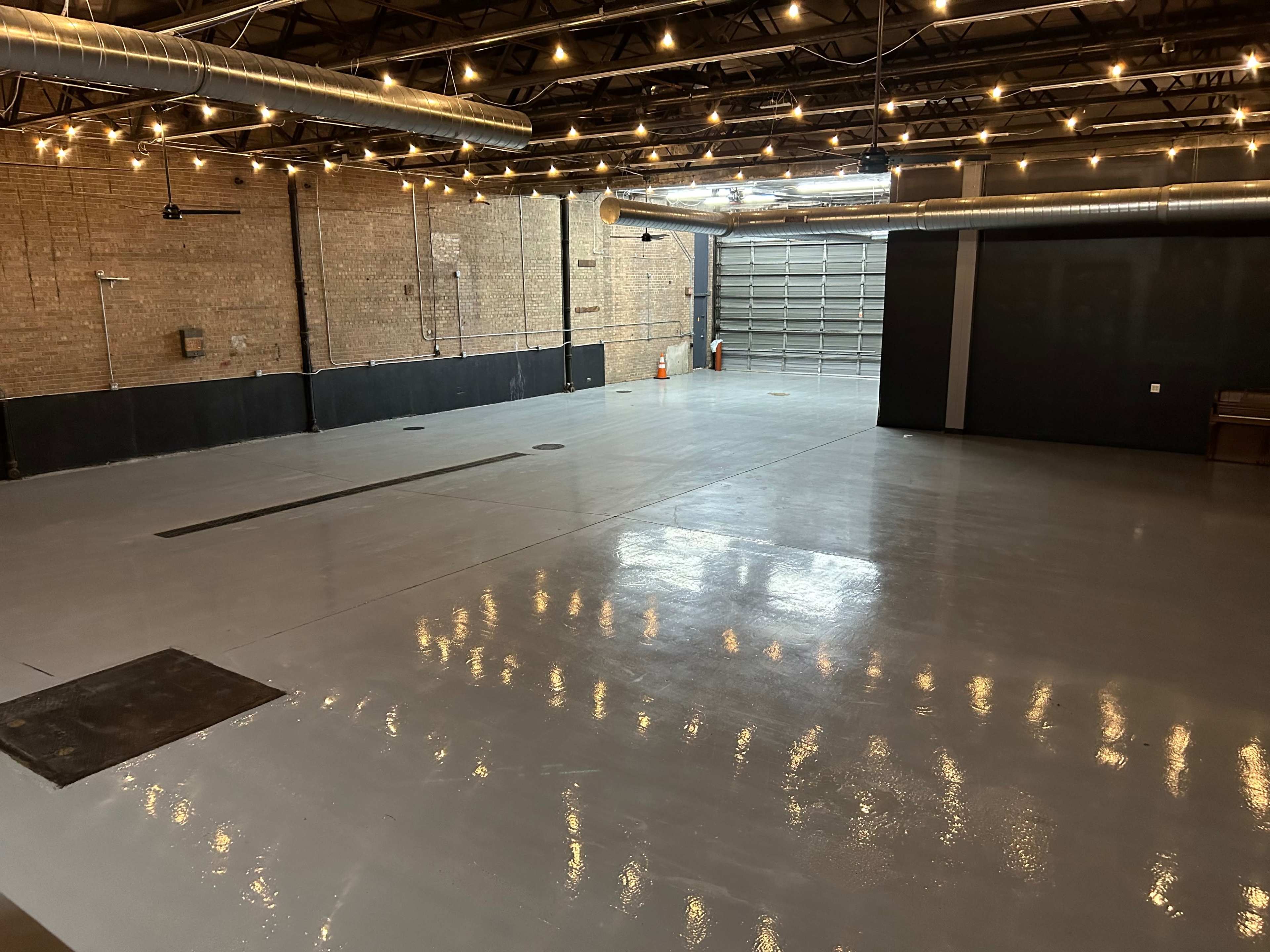 A spacious, industrial-style room with a polished concrete floor and exposed brick walls, featuring overhead lighting and a large garage door at one end.