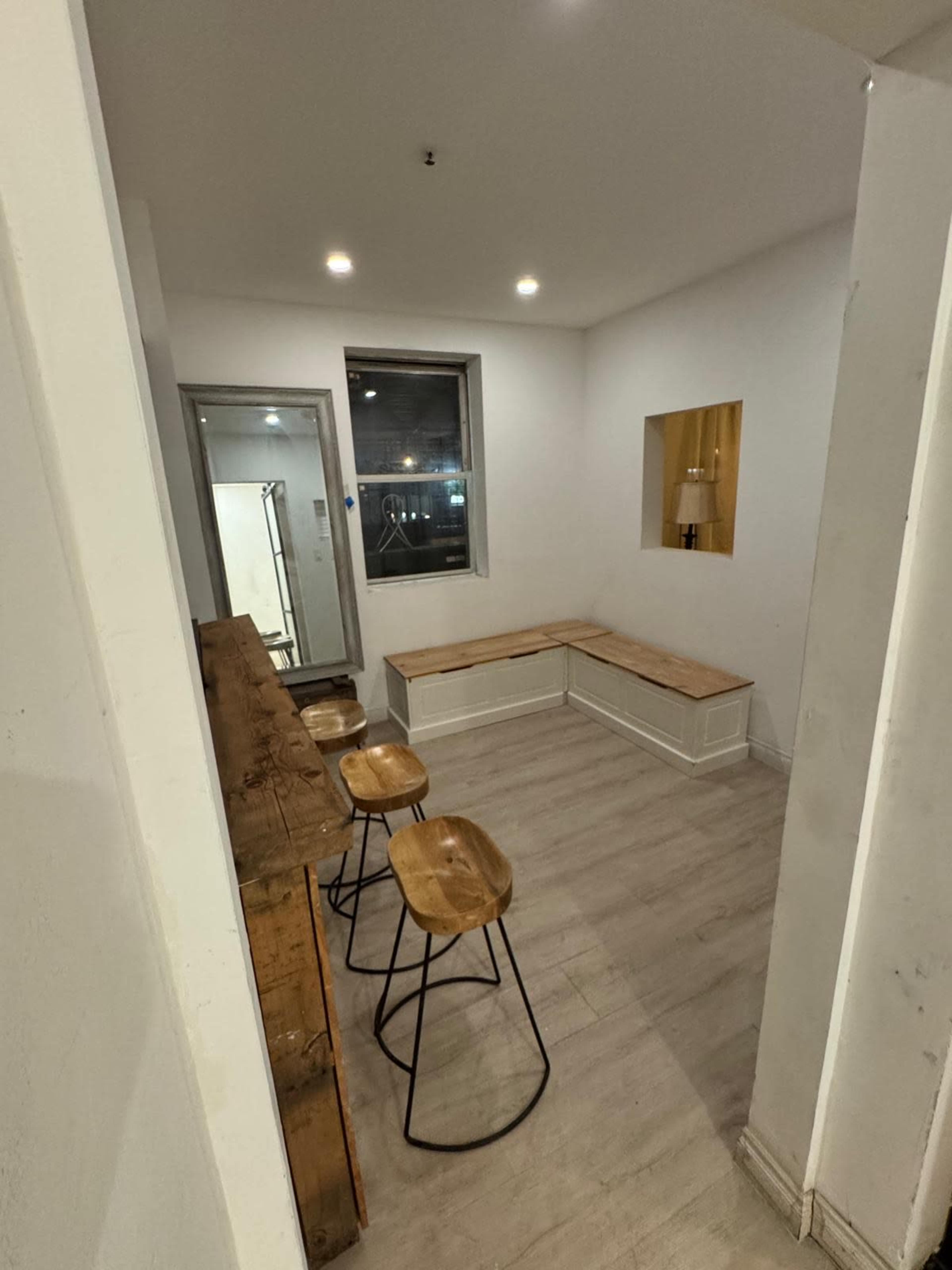 The image shows a small room with a wooden bench, a wooden table, and three stools, illuminated by overhead lights.