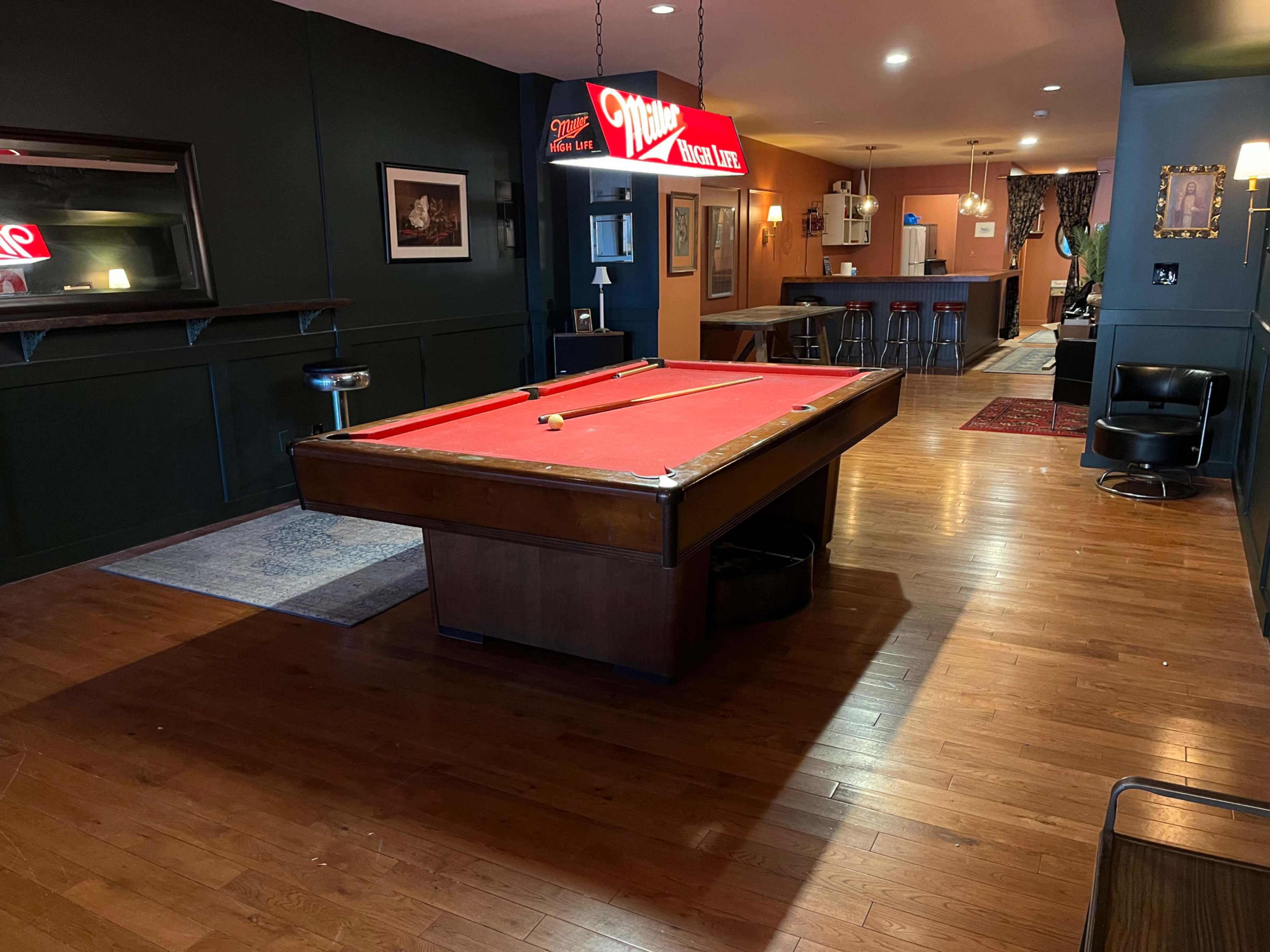 The image shows a game room featuring a red felt pool table at the center, surrounded by dark wood paneling and a bar area in the background.