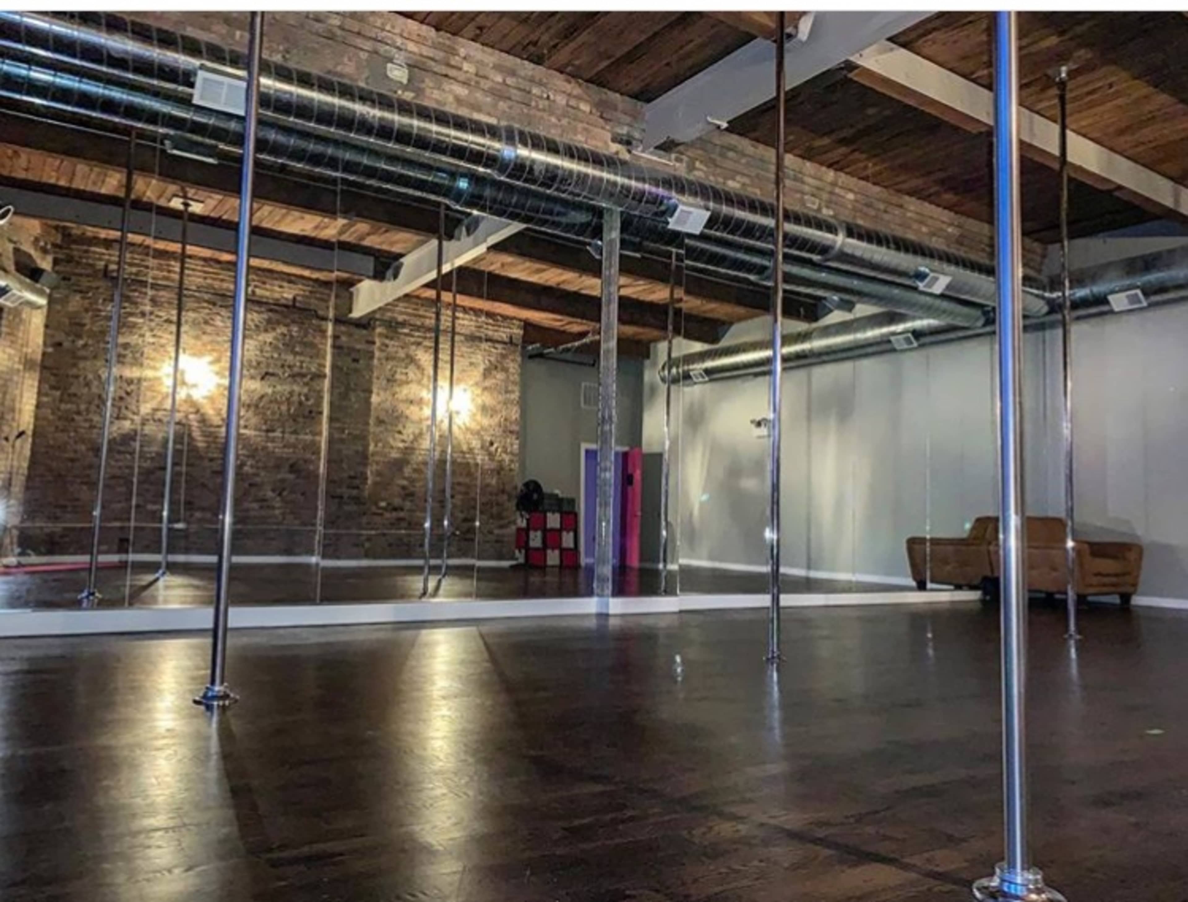 An empty dance studio featuring polished wooden floors, mirrored walls, and several pole dance installations.