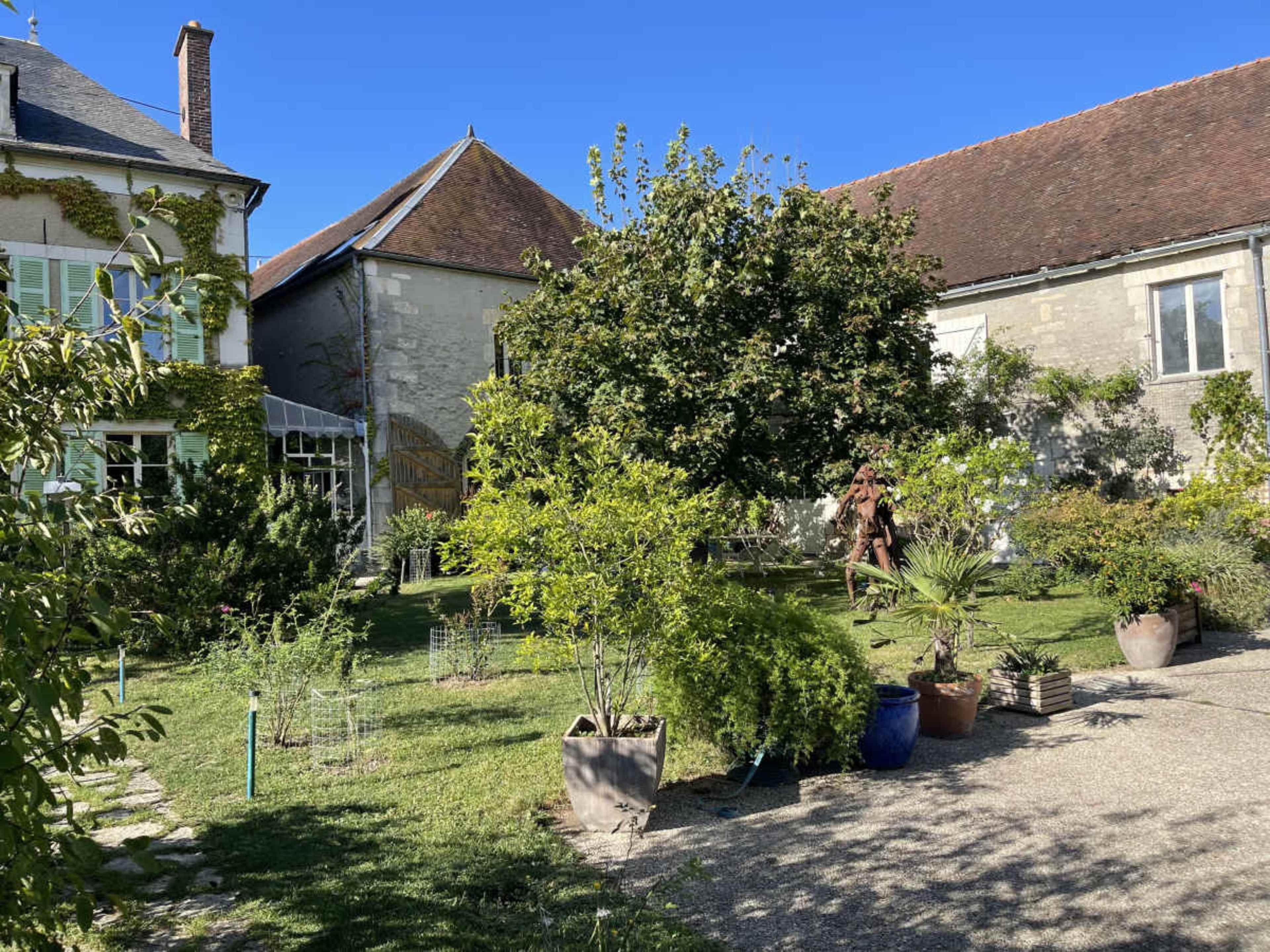 The image shows a garden with various plants and trees, surrounded by buildings with sloped roofs under a clear blue sky.