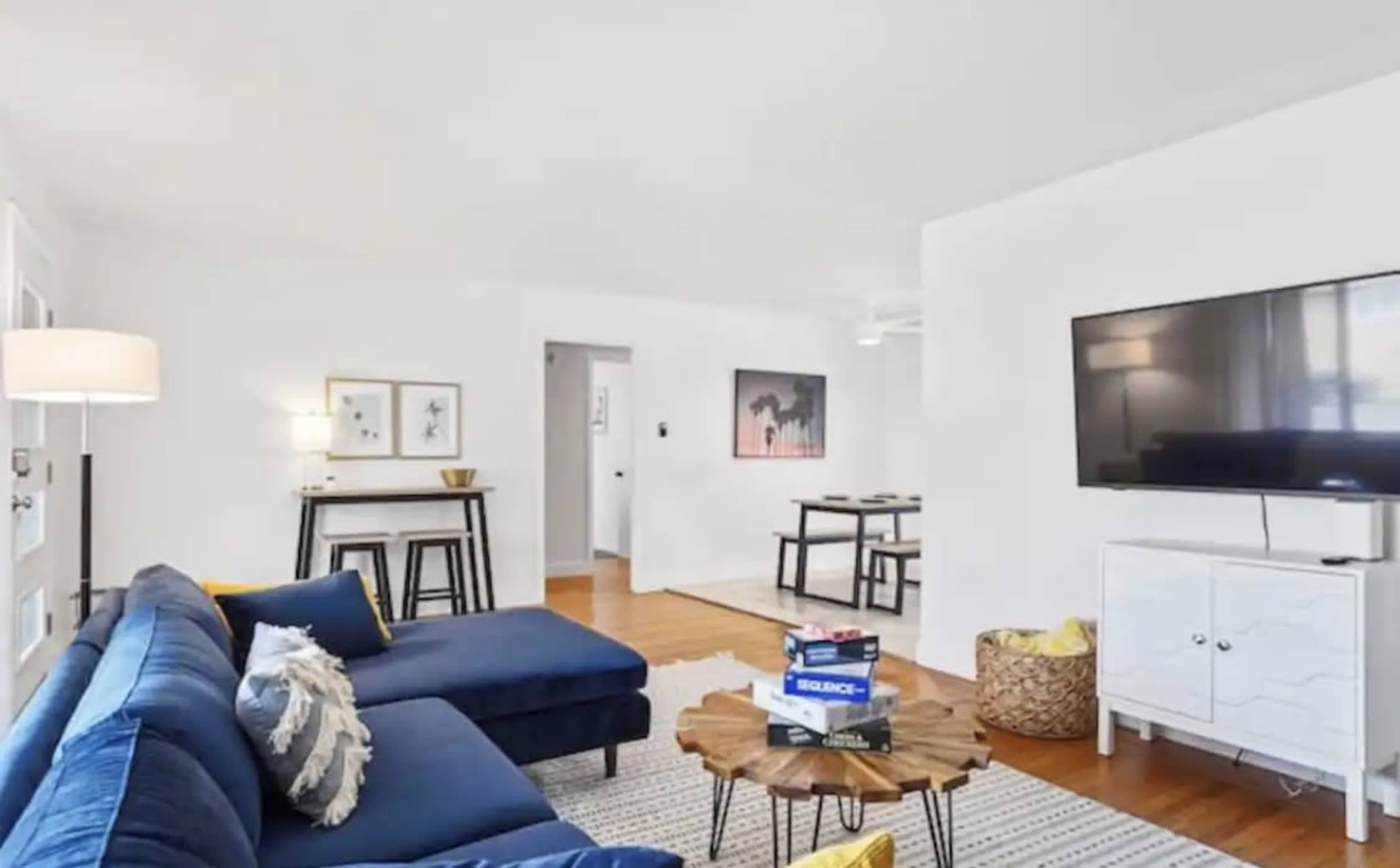 A brightly lit living room with a blue sectional sofa, a wooden coffee table, a television on a white cabinet, and a dining area in the background.