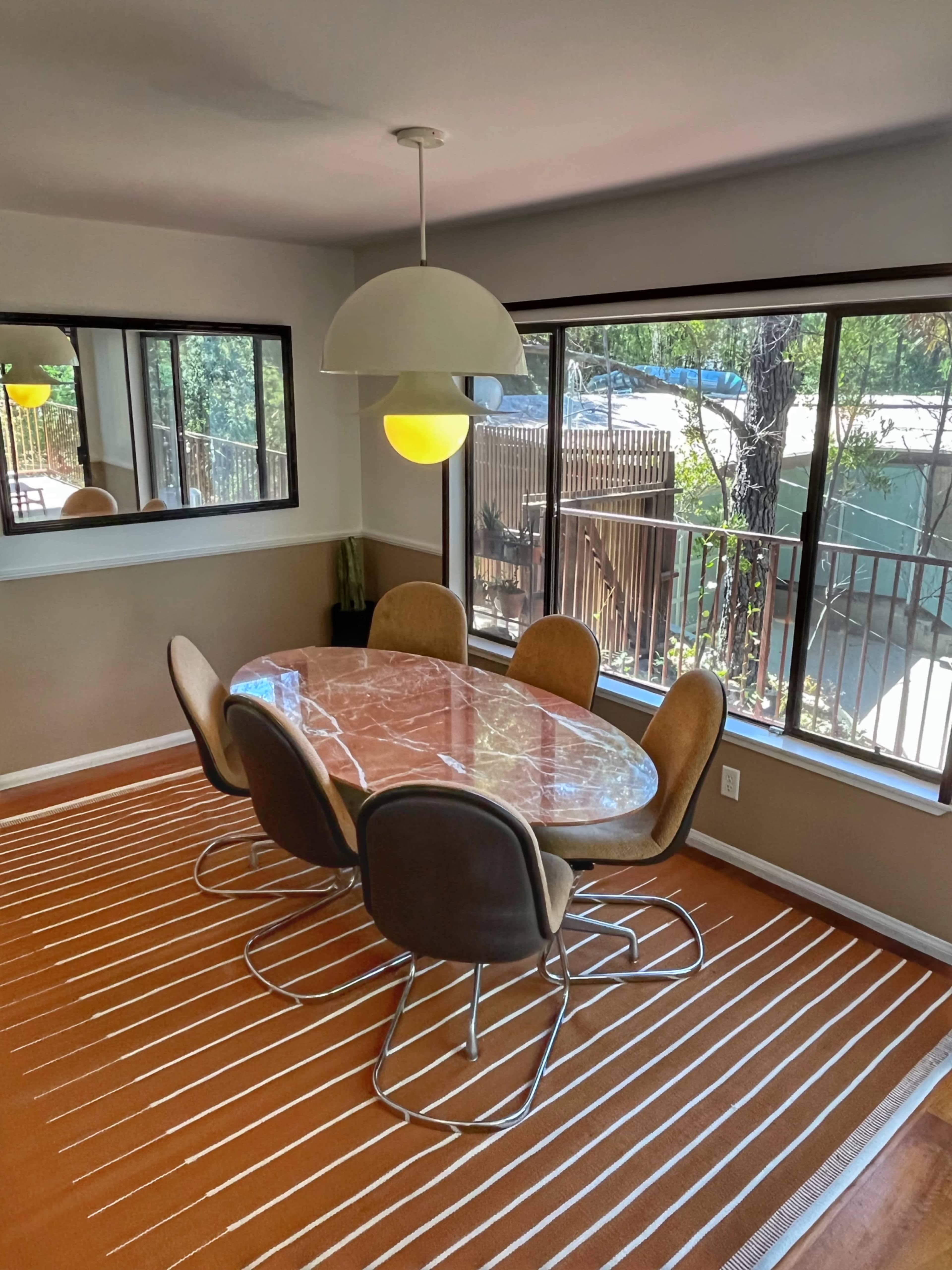 A modern dining area features an oval marble table surrounded by four upholstered chairs, with a pendant light overhead and large windows overlooking a wooden deck.