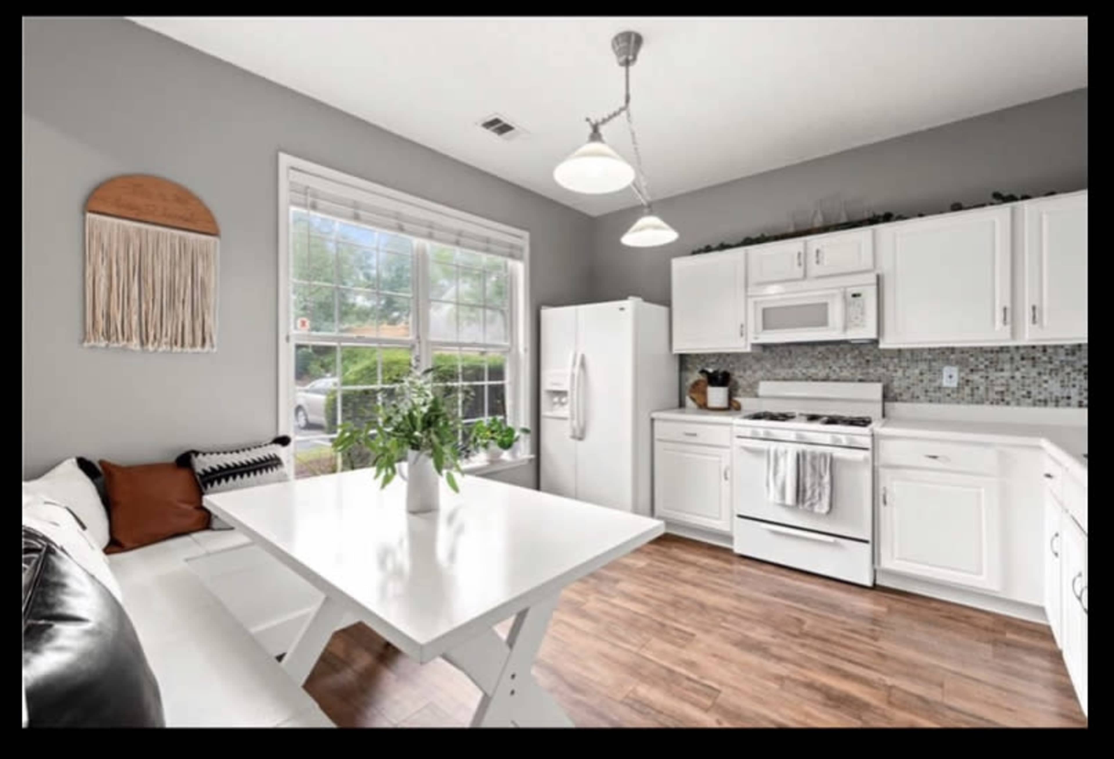 A kitchen features white cabinets, a large window, and a dining table with a bench, illuminated by pendant lights.