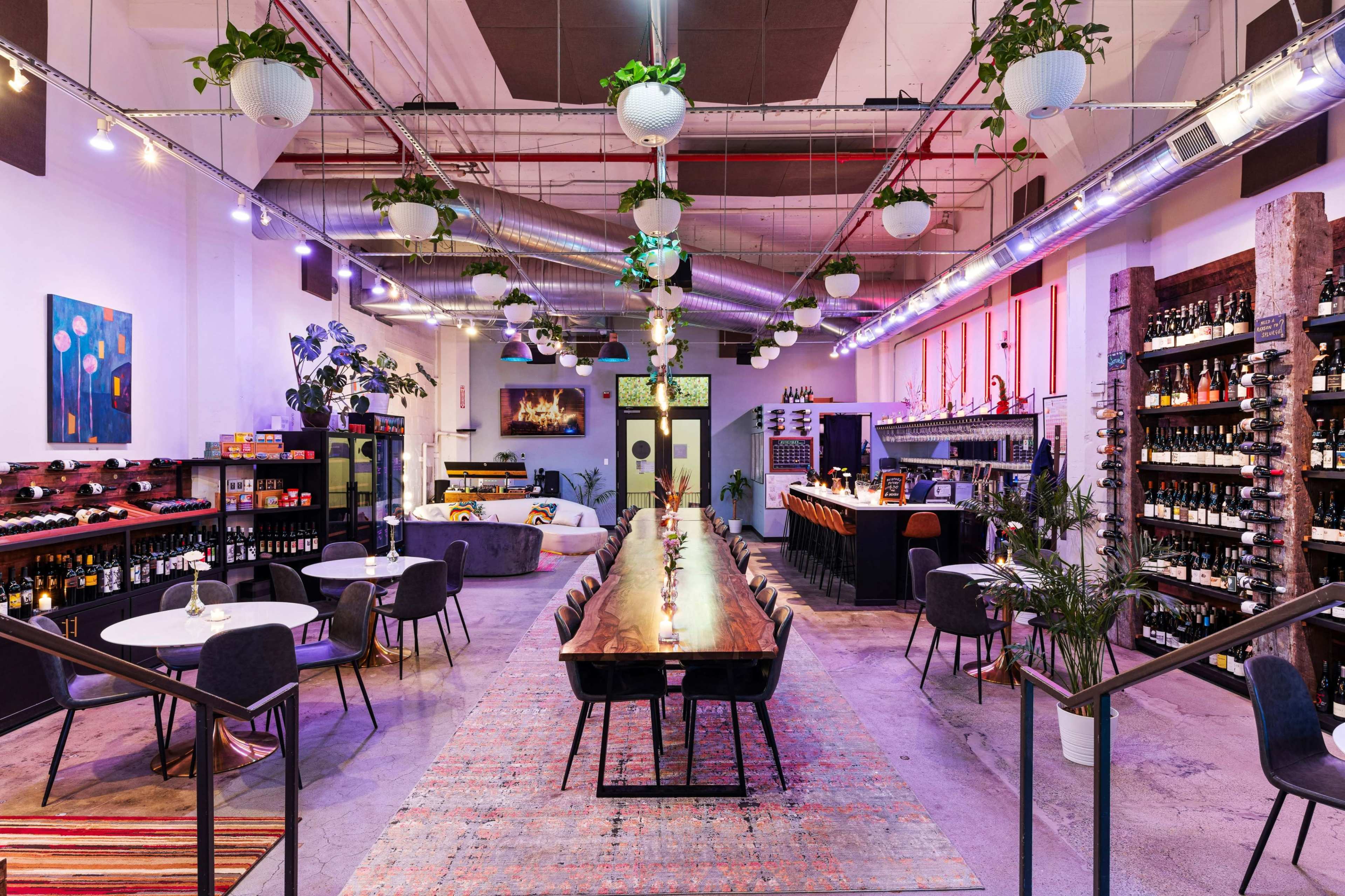 A modern, spacious restaurant with a long wooden dining table, surrounded by plants, shelves of wine, and various seating arrangements.