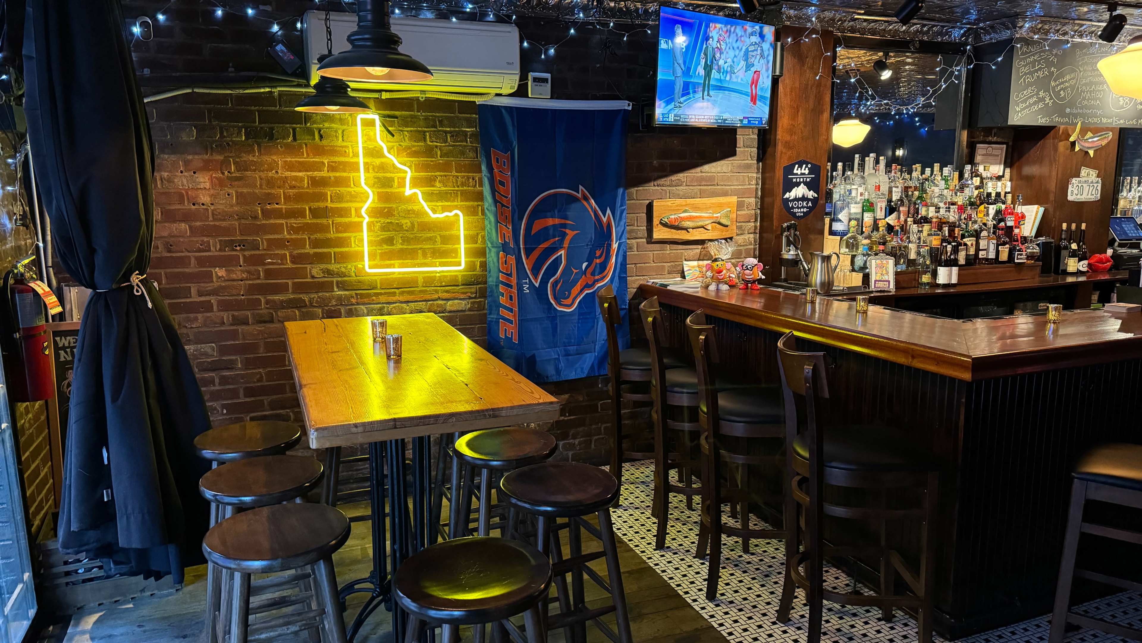 A cozy bar area with a wooden table, several tall chairs, and a bright neon outline of the state of Idaho on the wall, alongside a Boise State flag and a well-stocked bar.