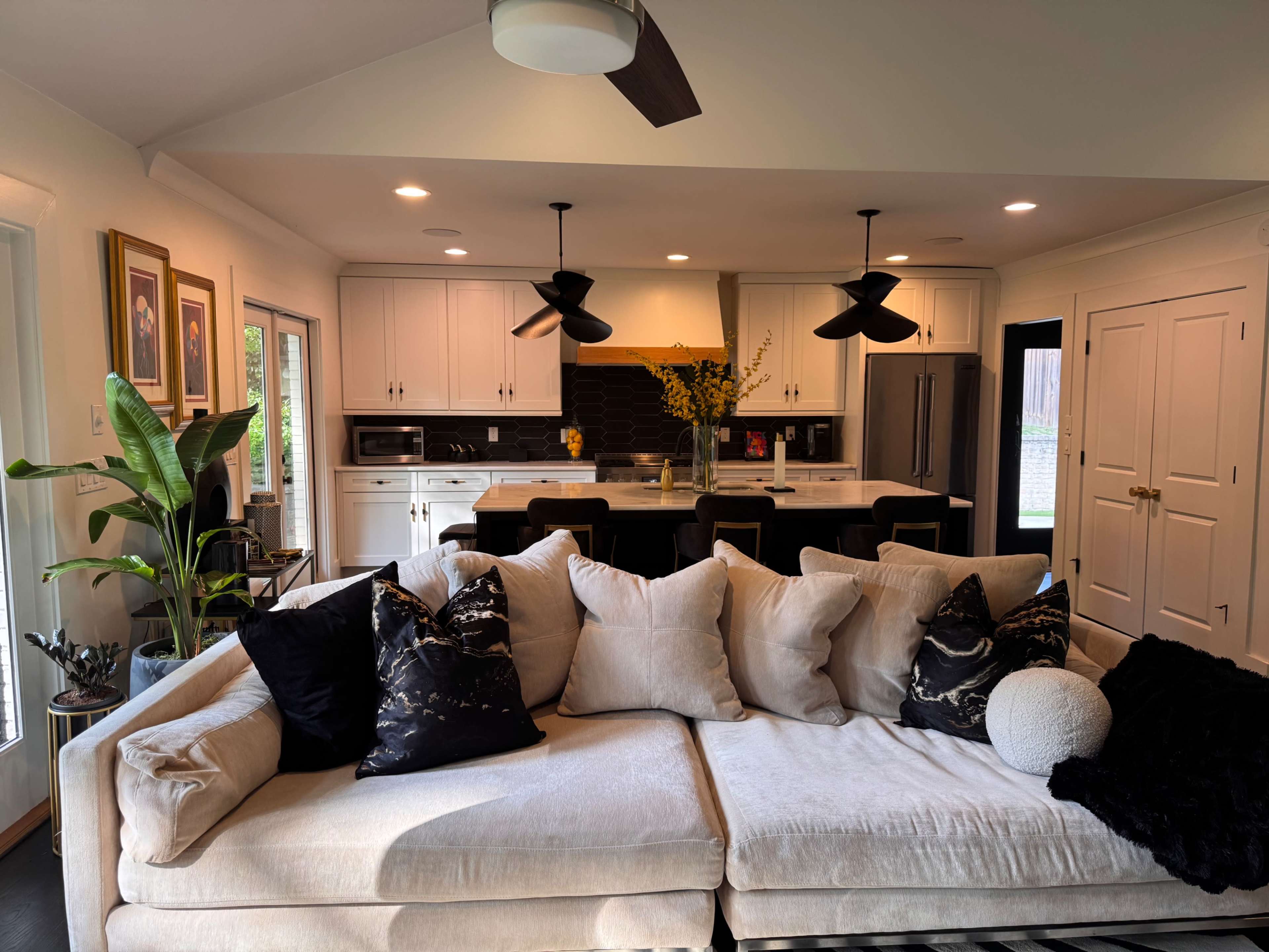 The image shows a modern living room with a light-colored sofa positioned in front of a kitchen featuring dark cabinetry and a dining area with black bar stools.