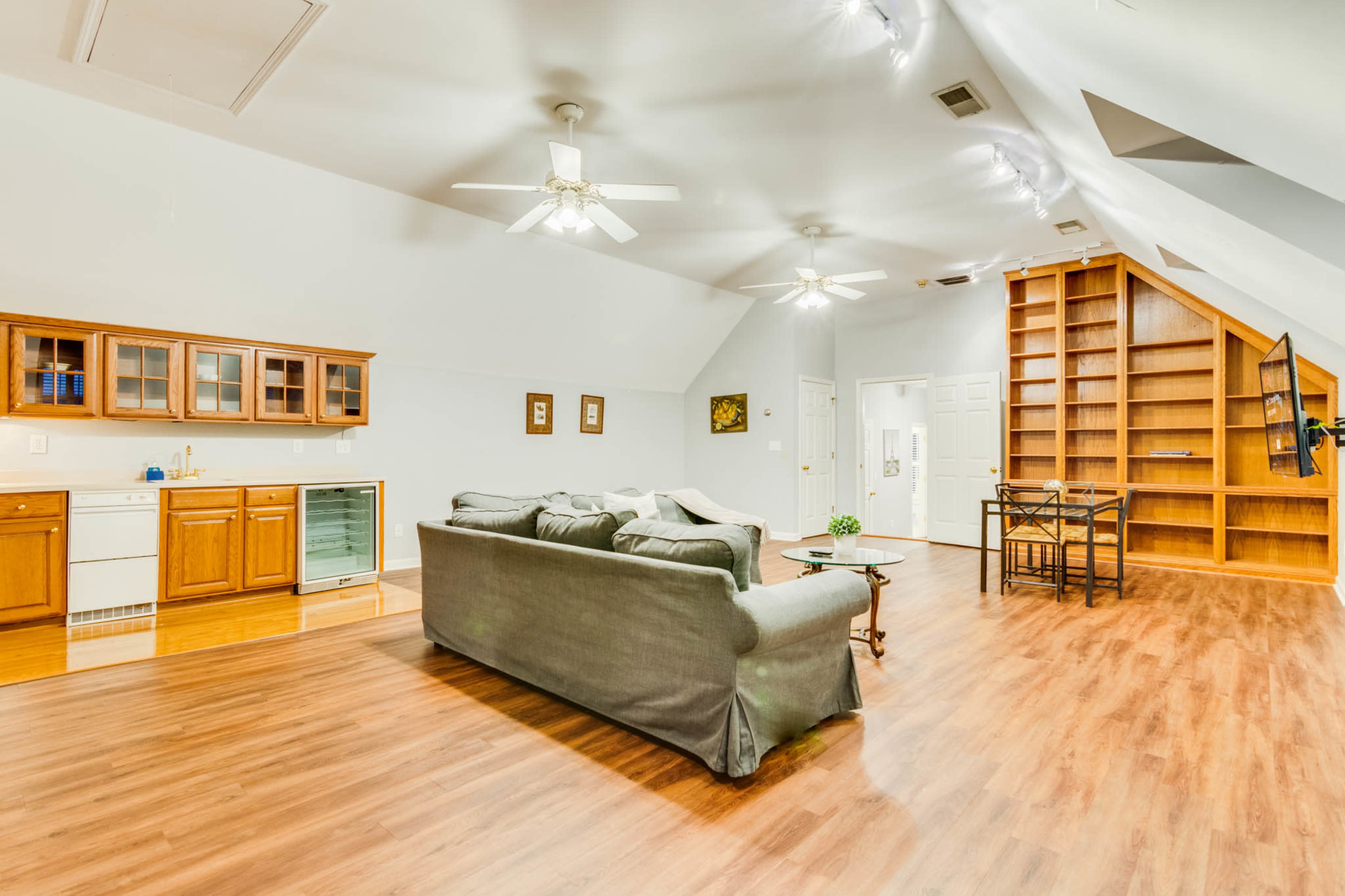 The image shows a spacious attic room with a living area featuring a gray sofa, a kitchenette with wooden cabinets, a dining table, and a built-in bookshelf.