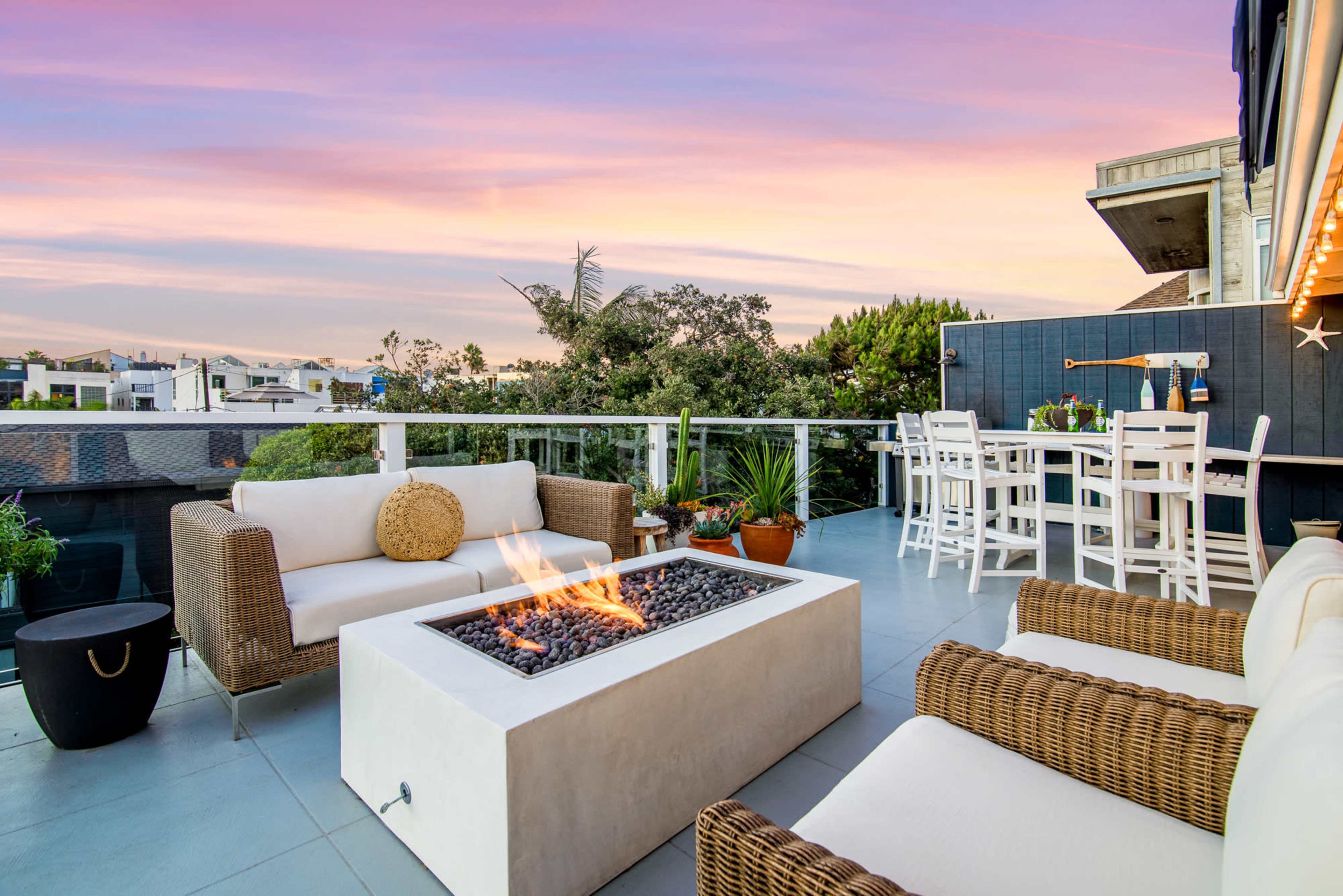 A modern outdoor patio features two wicker sofas surrounding a fire pit, with a dining area and vibrant sunset in the background.