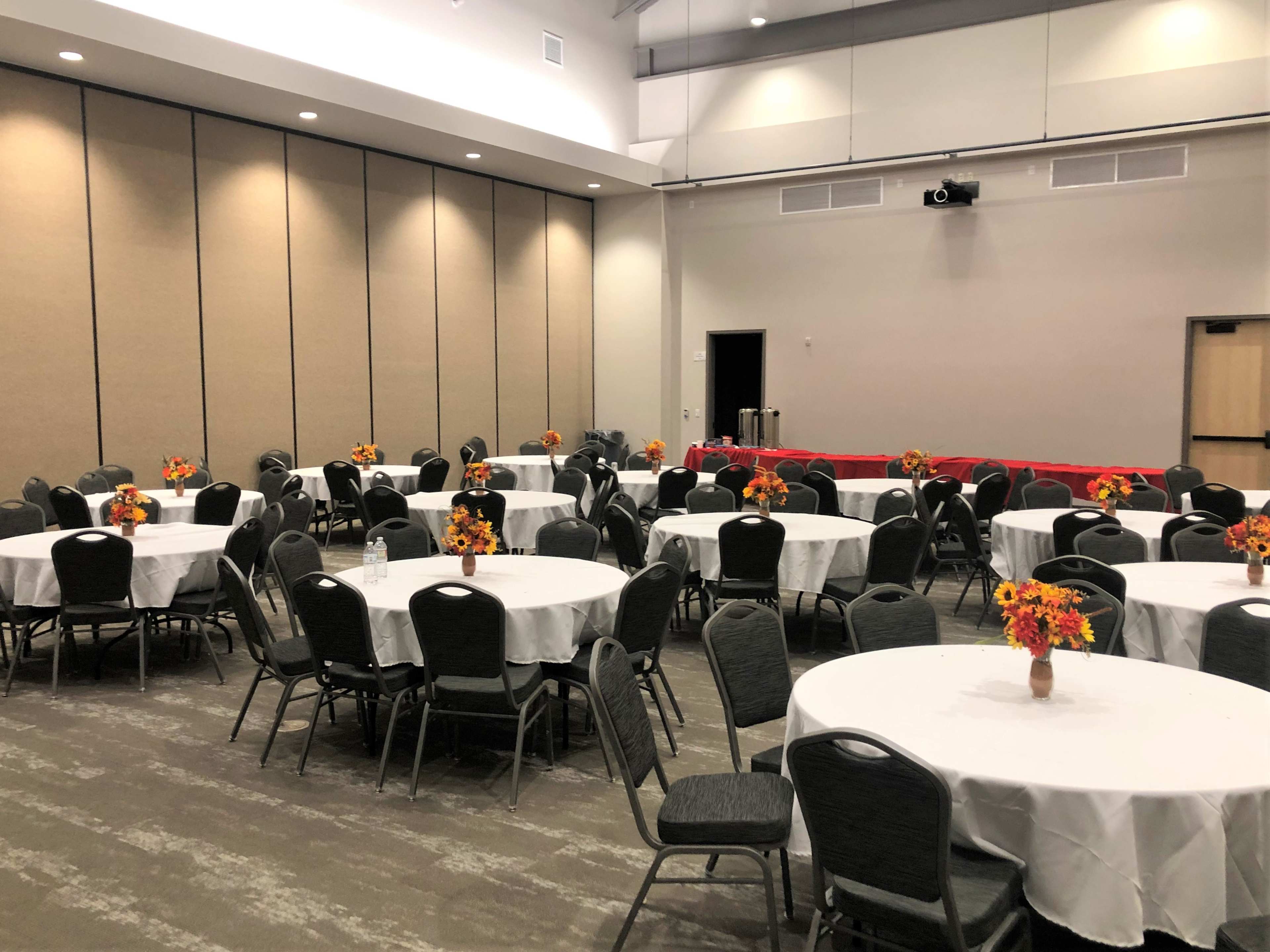 A large meeting room with round tables covered in white tablecloths and decorated with flower centerpieces, arranged in a grid pattern.