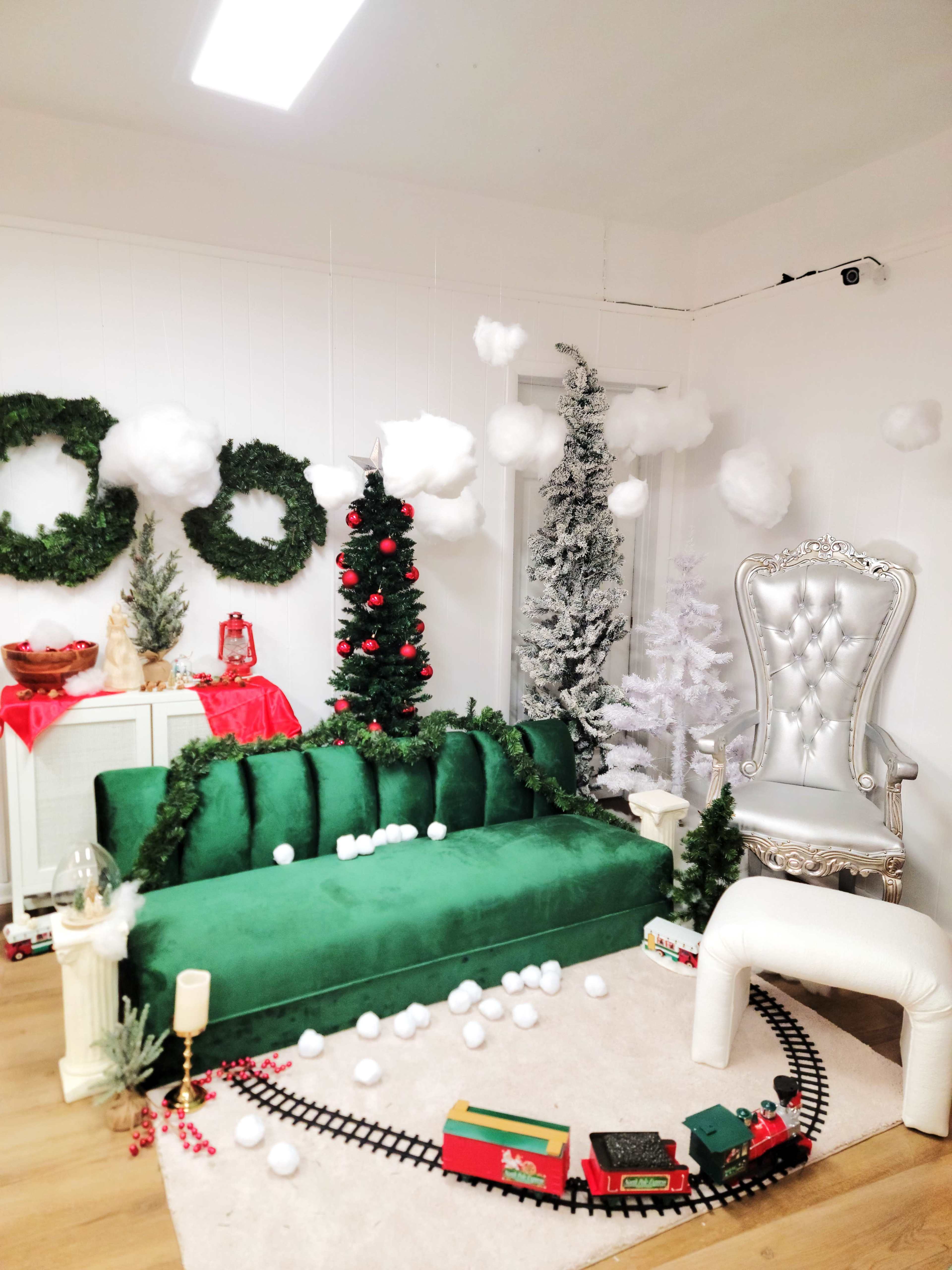 The scene features a decorated room with a green velvet couch, two Christmas trees, white fluffy decorations, and a train set on the floor.