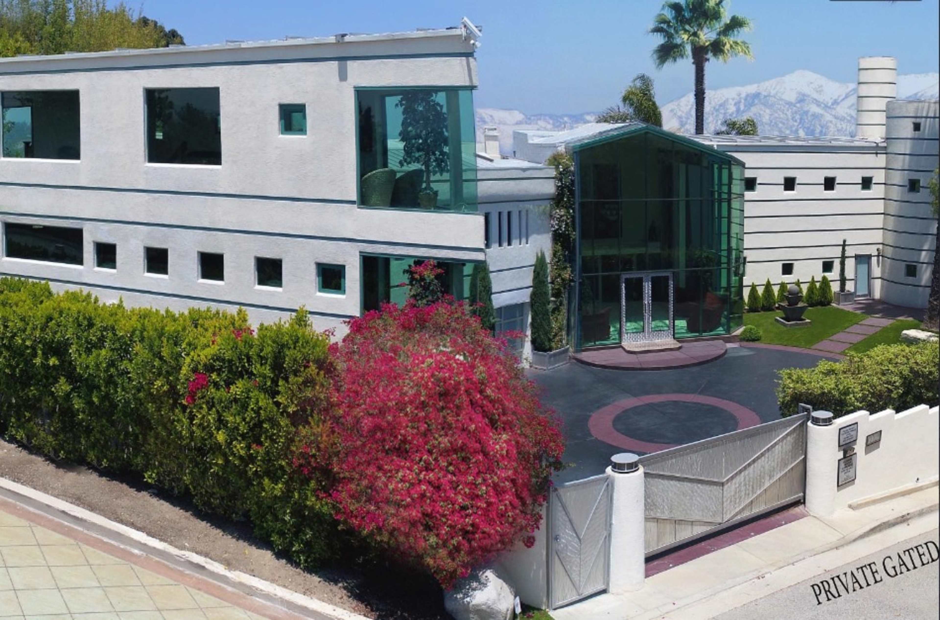 The image shows a modern, multi-story building with large windows and a landscaped entrance, bordered by a vibrant flowering shrub and a gate labeled "PRIVATE GATED."