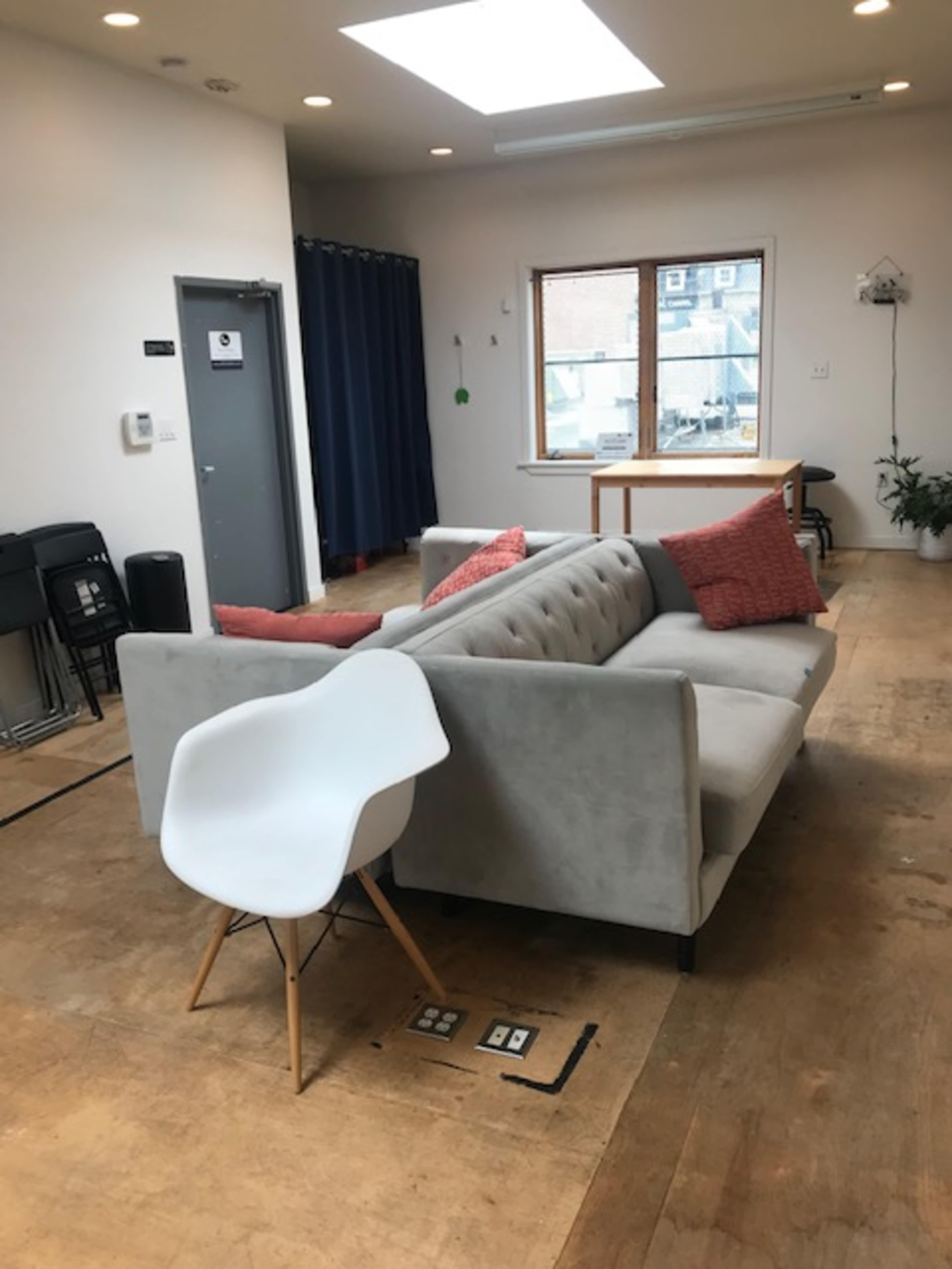 A gray sofa with pillows is positioned in a room featuring wooden flooring, a large window, and a white chair nearby.