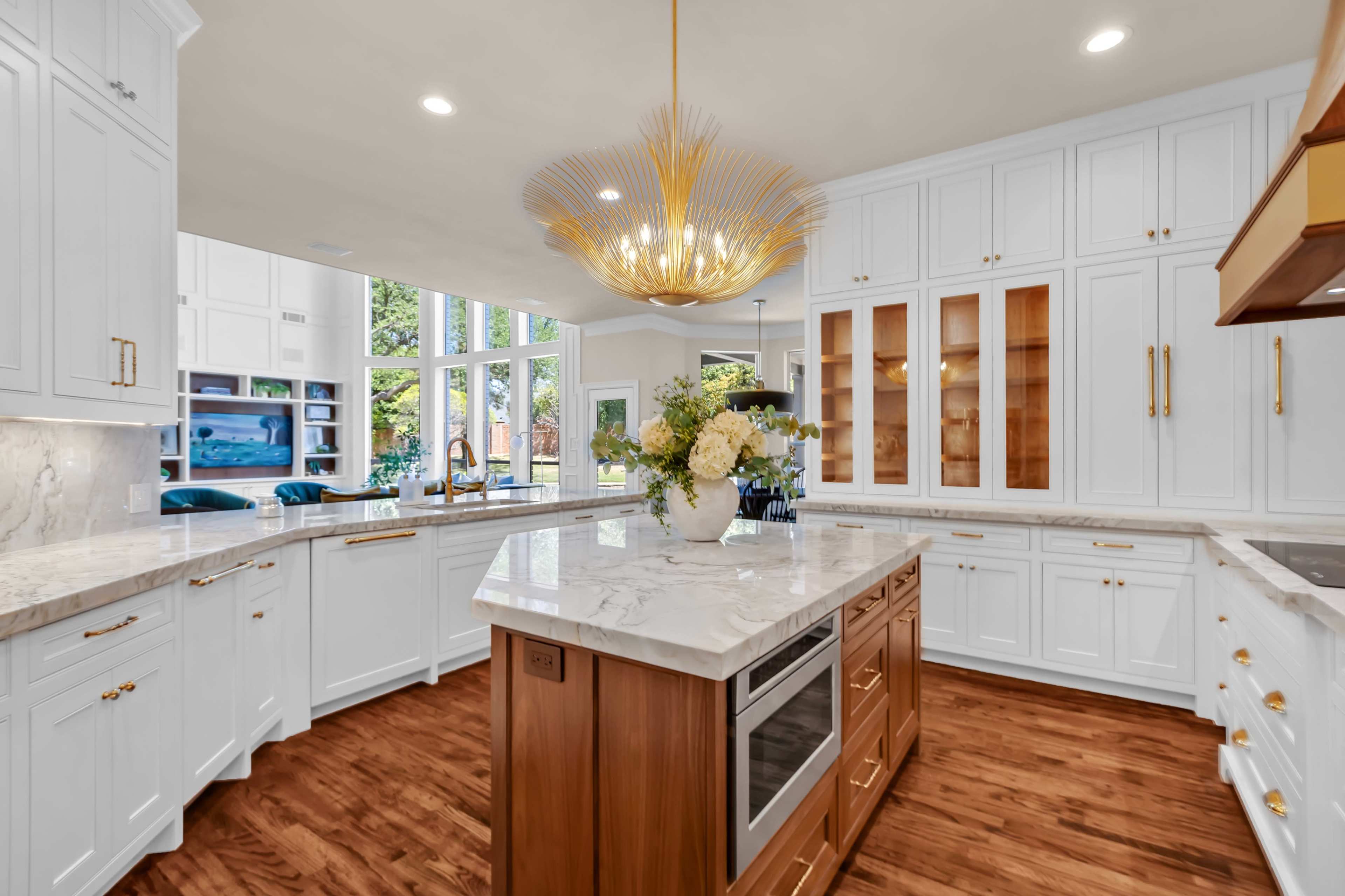 The image shows a modern kitchen featuring a central island with a marble countertop, white cabinetry, and large windows that allow natural light to fill the space.