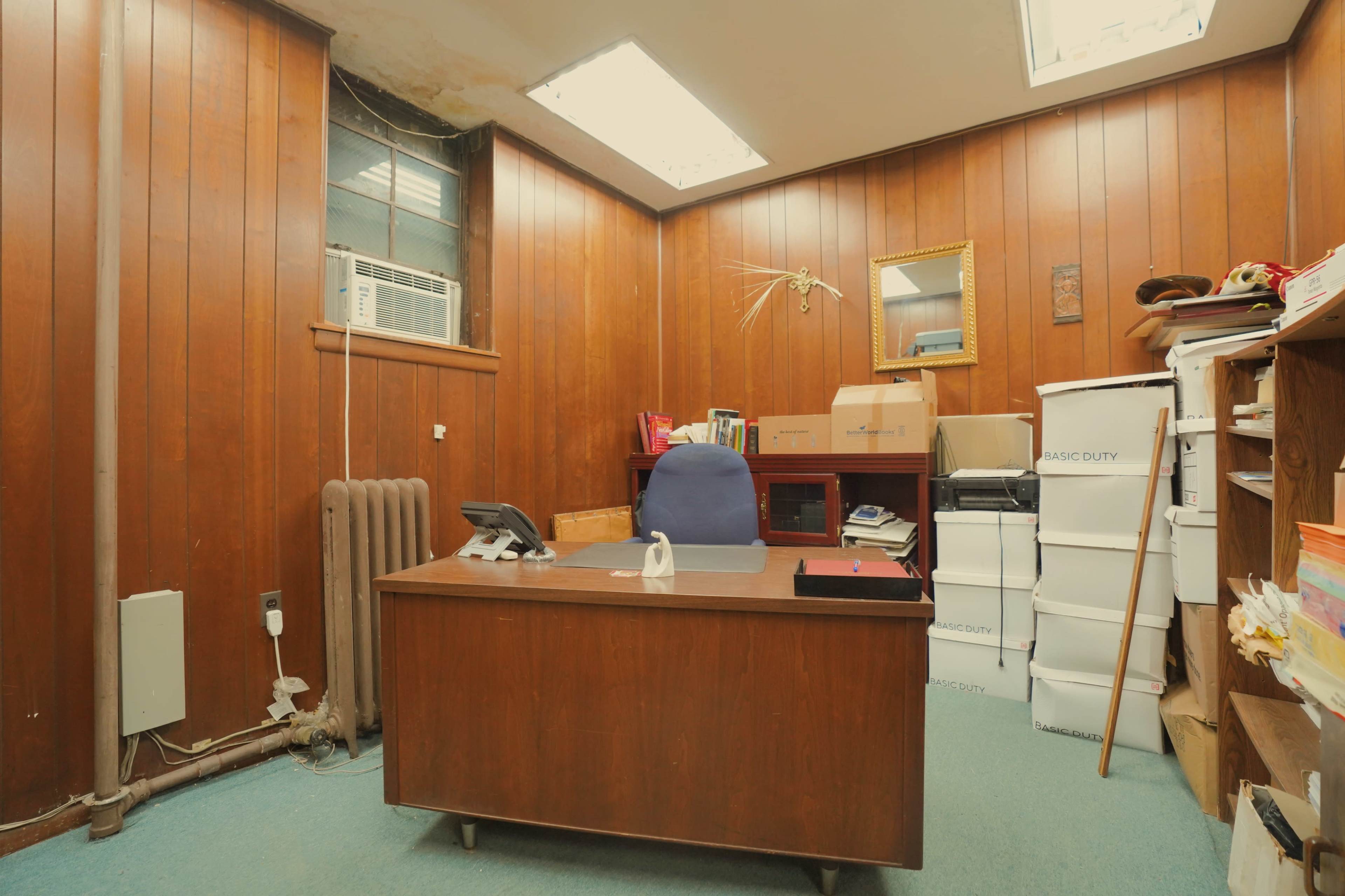 The image shows a small office with wooden paneling, a desk with a telephone, and boxes stacked on the floor and shelves.