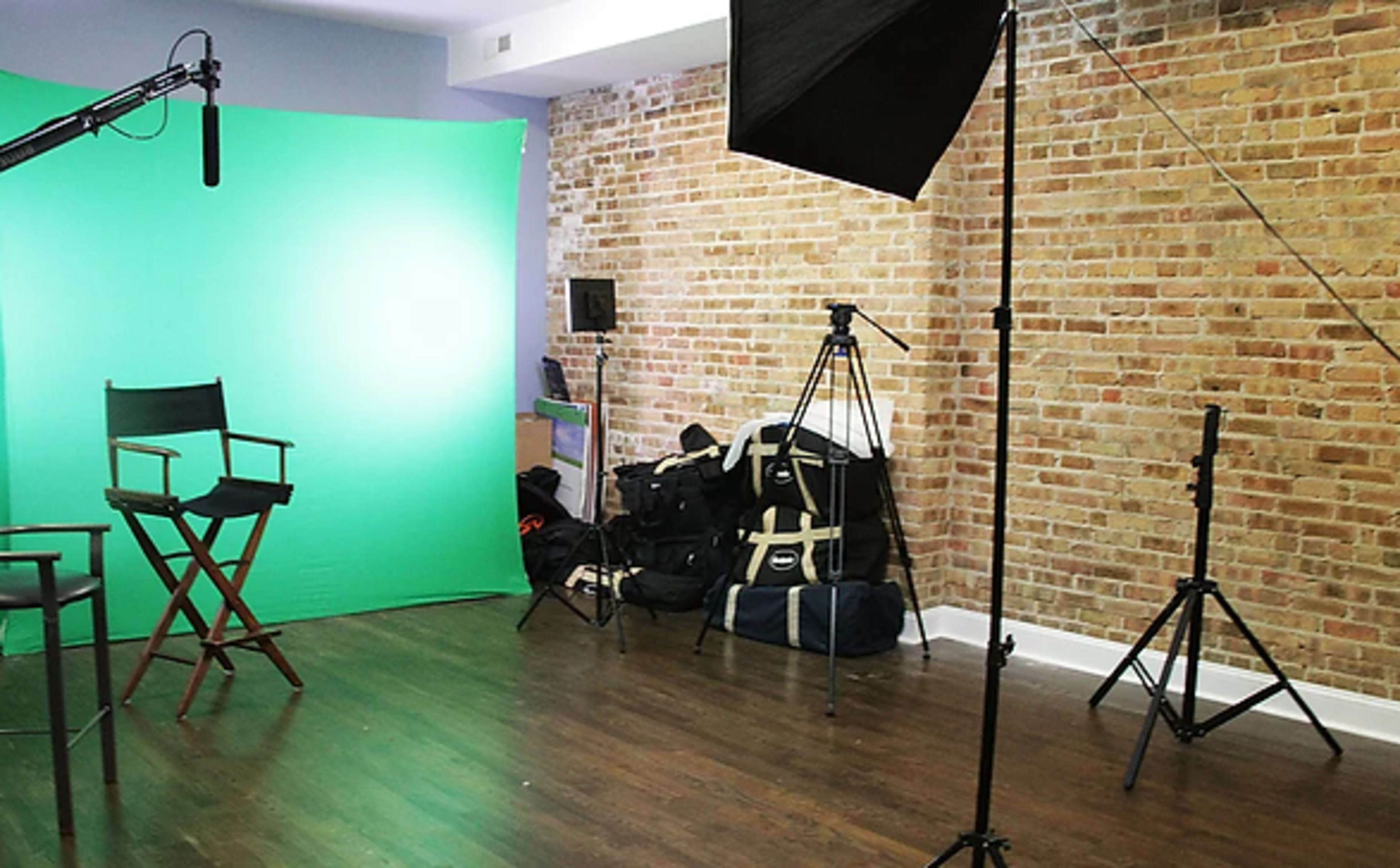 A video production setup featuring a green screen, director's chair, lighting equipment, and tripods in a studio with exposed brick walls.