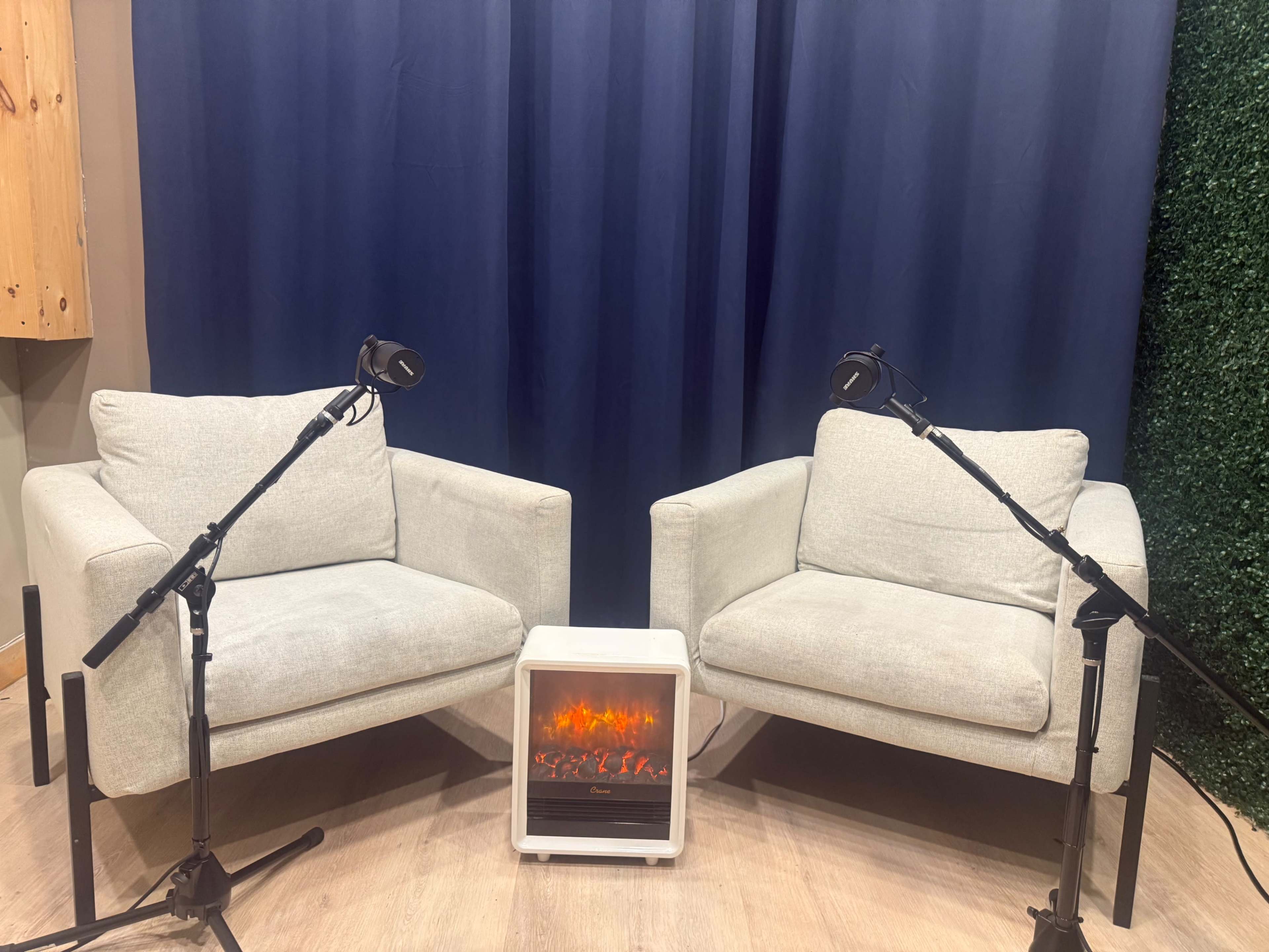 Two light gray chairs are positioned facing each other, with a small electric fireplace between them and black microphones attached to stands aiming towards the seats, all set against a dark blue backdrop.