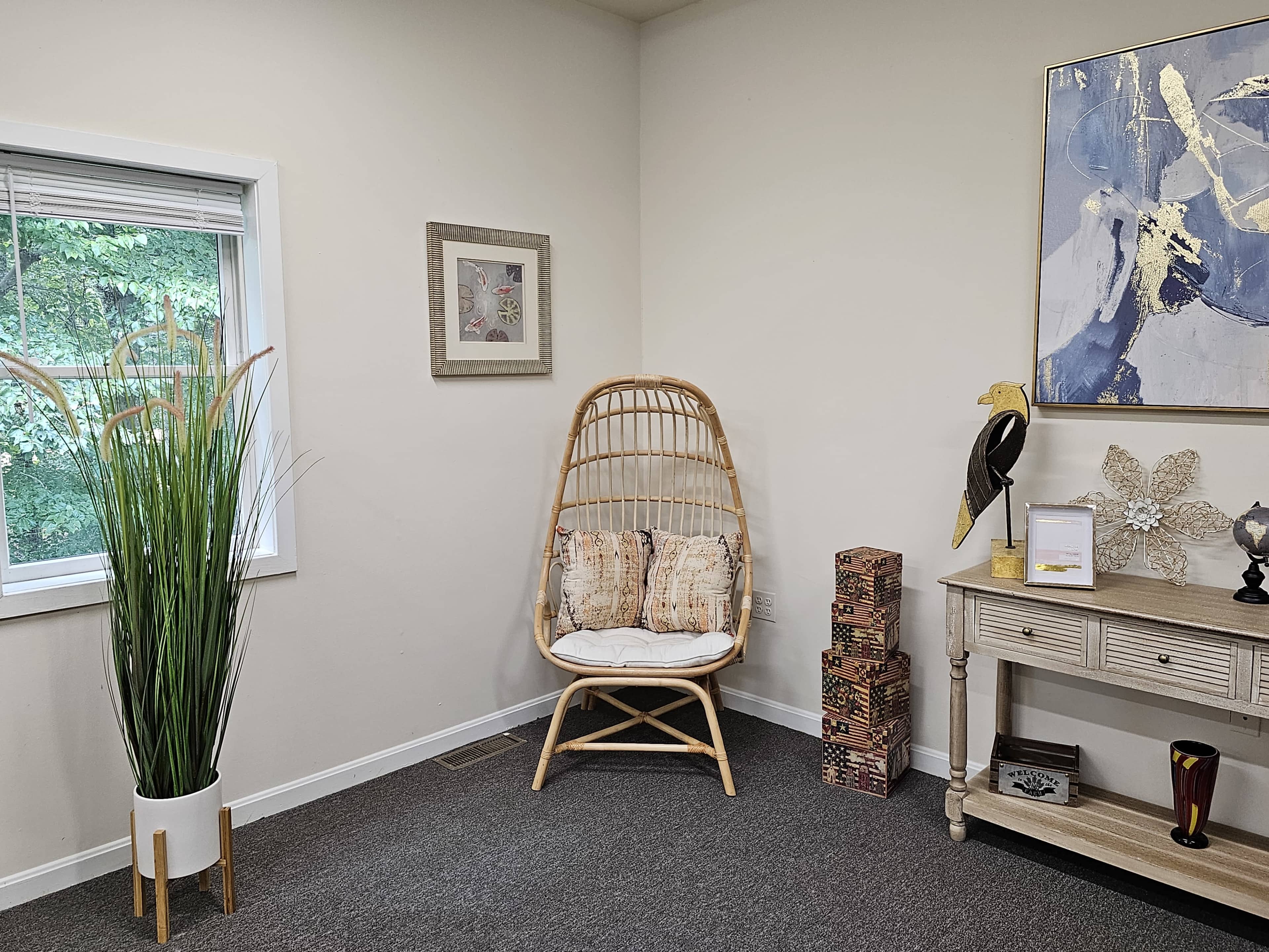 A cozy corner features a rattan chair with cushions beside a window, accompanied by decorative plants and a small table adorned with various items.