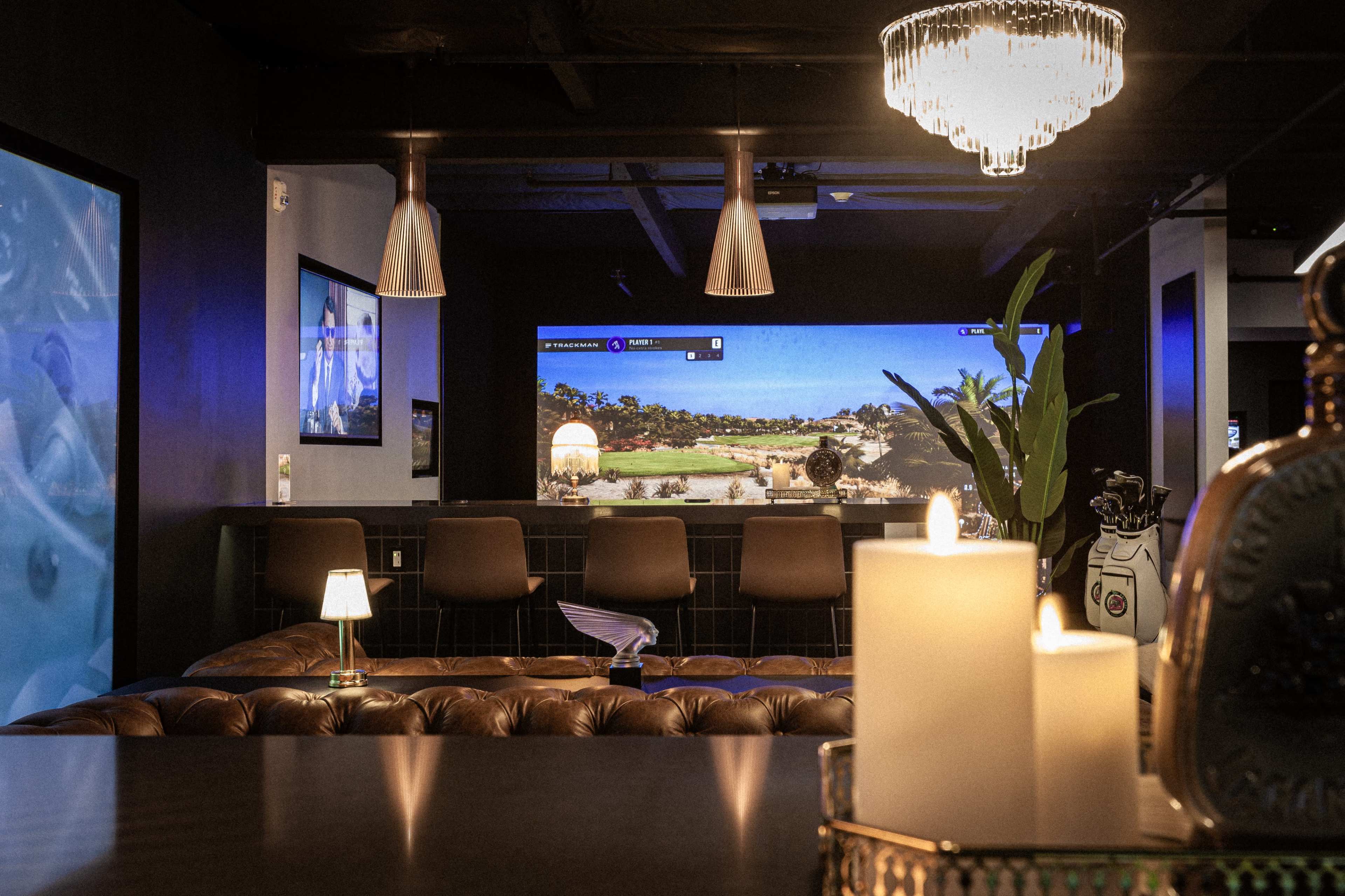 The scene features a stylish lounge area with a bar, illuminated by pendant lights, a large screen displaying a golf course, and decorative elements like candles and plants.