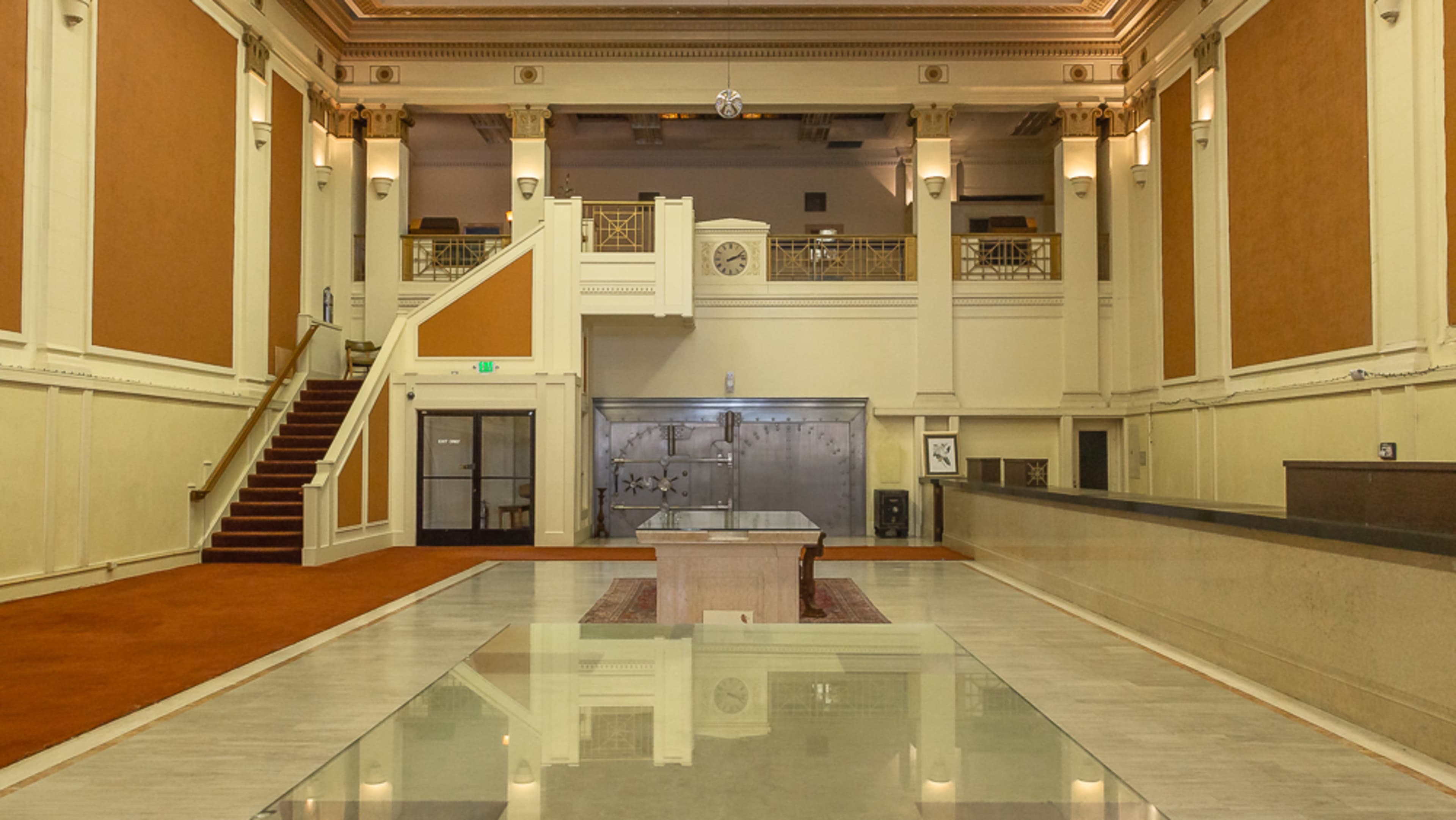 An empty grand hall with high ceilings, a stairway on the left, and a large vault door in the background.