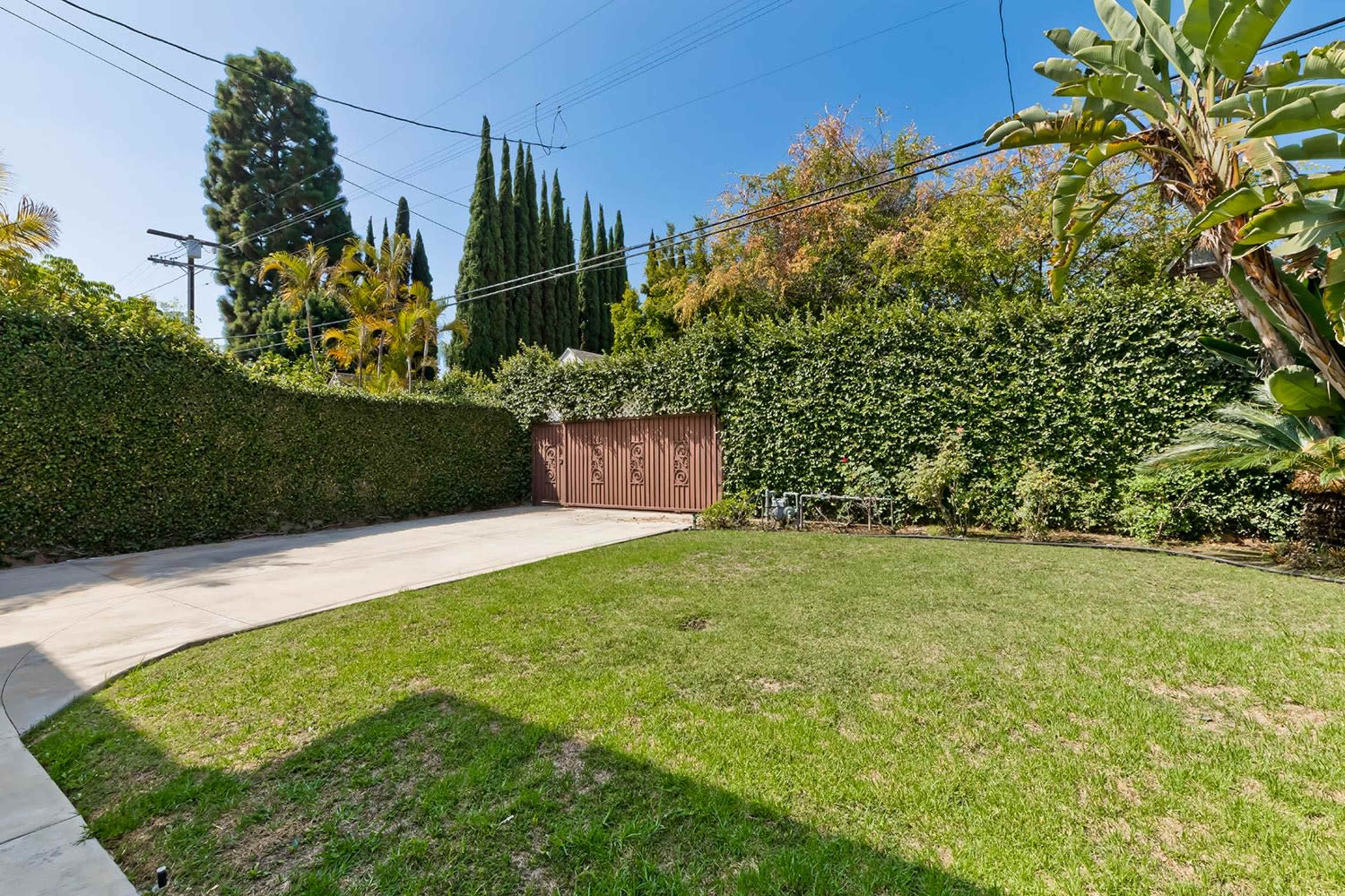 The image shows a spacious, well-maintained backyard with a concrete area, green grass, and tall hedges surrounding the perimeter.