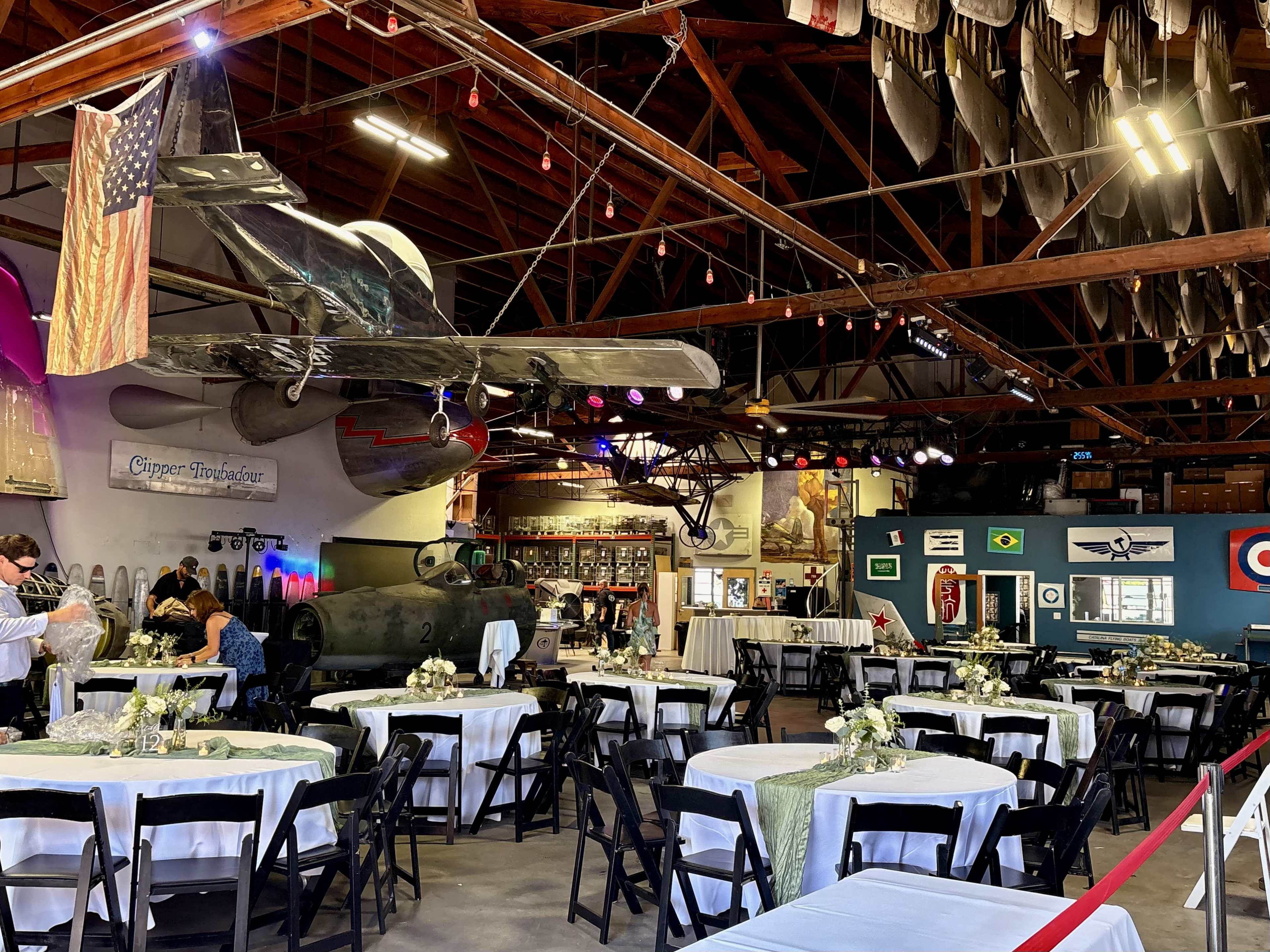 The interior of an event space features round tables with floral centerpieces, an airplane hanging from the ceiling, and various military artifacts displayed on the walls.