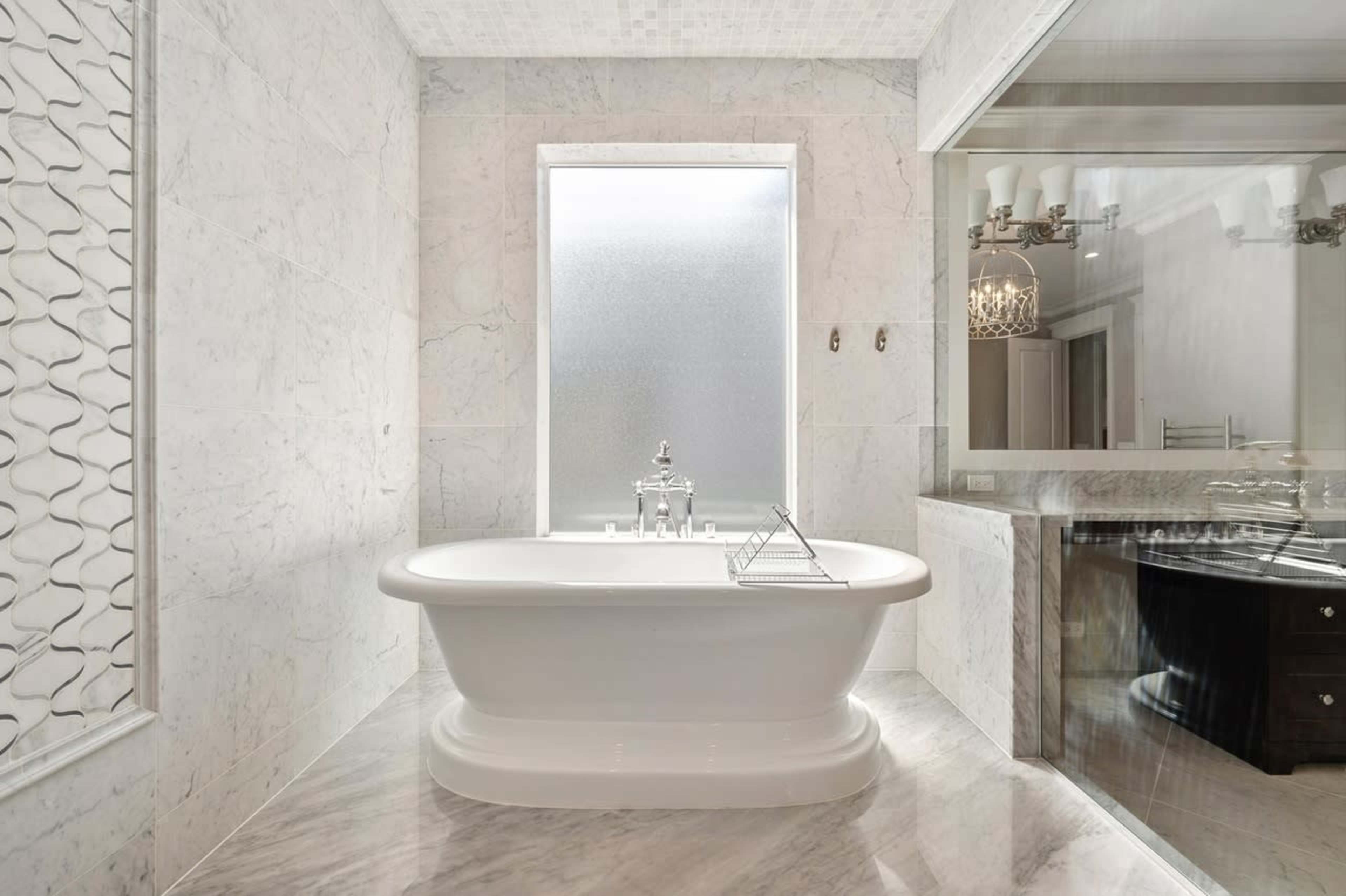 A modern bathroom featuring a freestanding white bathtub, marble walls, and a large window with frosted glass.