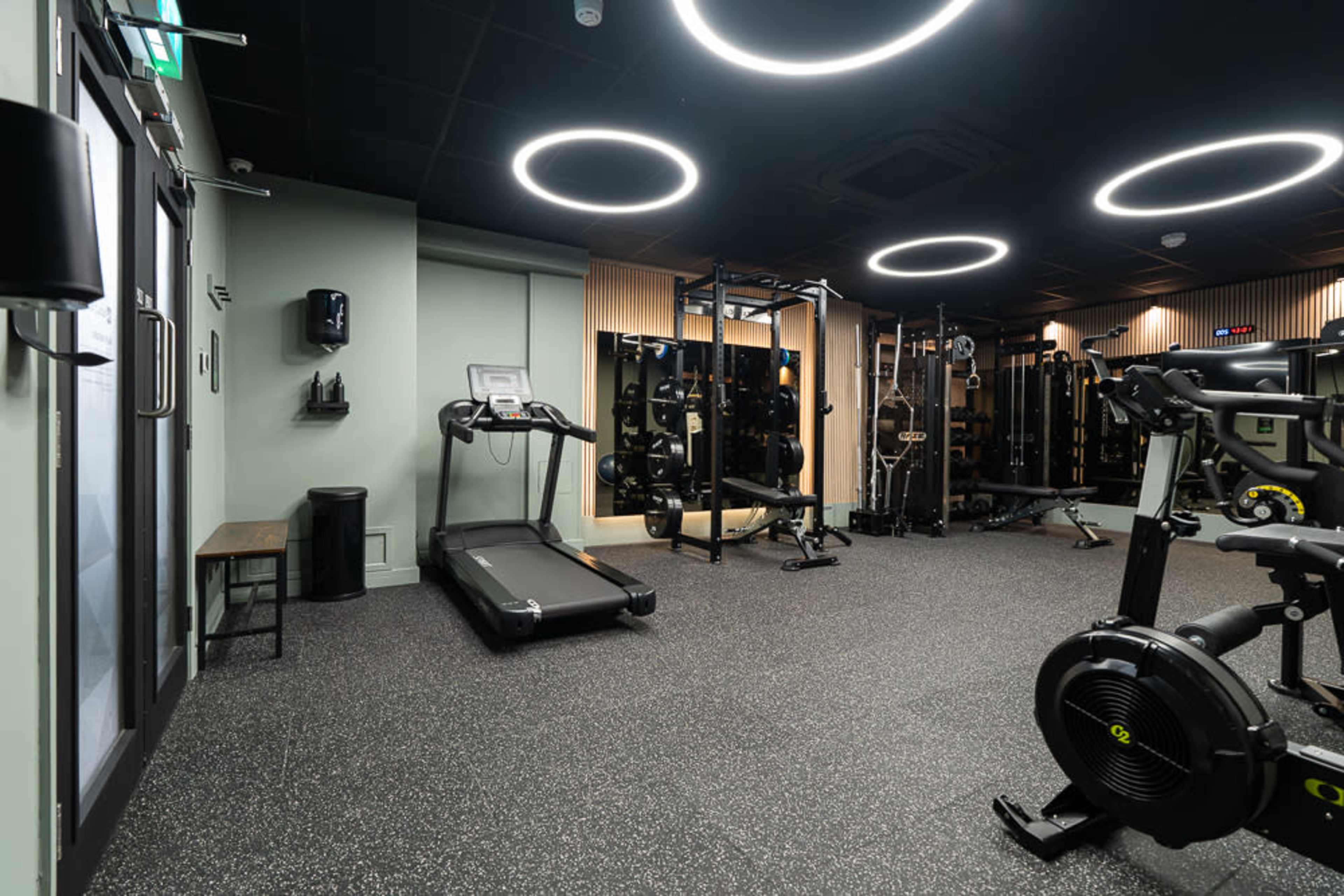 The image shows the interior of a modern gym featuring exercise equipment, including a treadmill and weightlifting stations, with circular overhead lights illuminating the space.