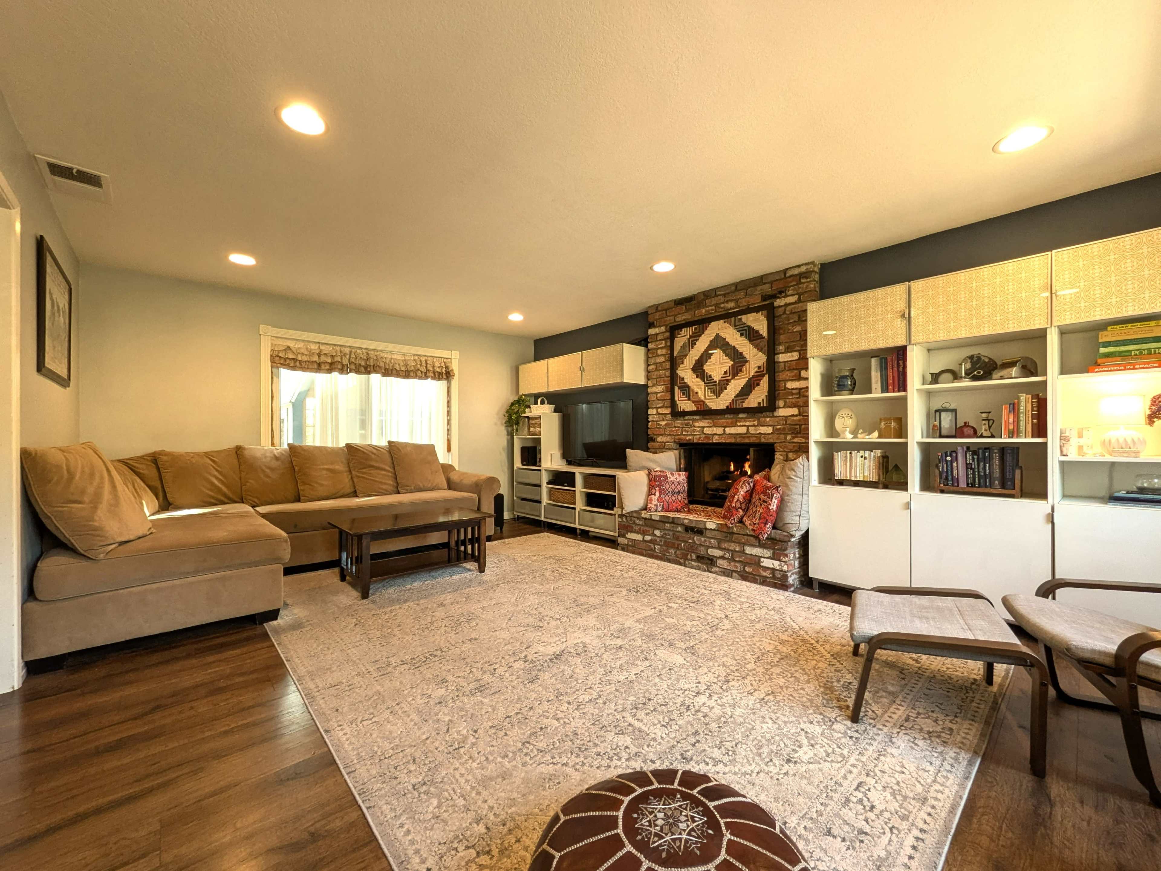 A cozy living room features a large sectional sofa, a brick fireplace with decorative accents, built-in shelves, and a light-colored area rug.