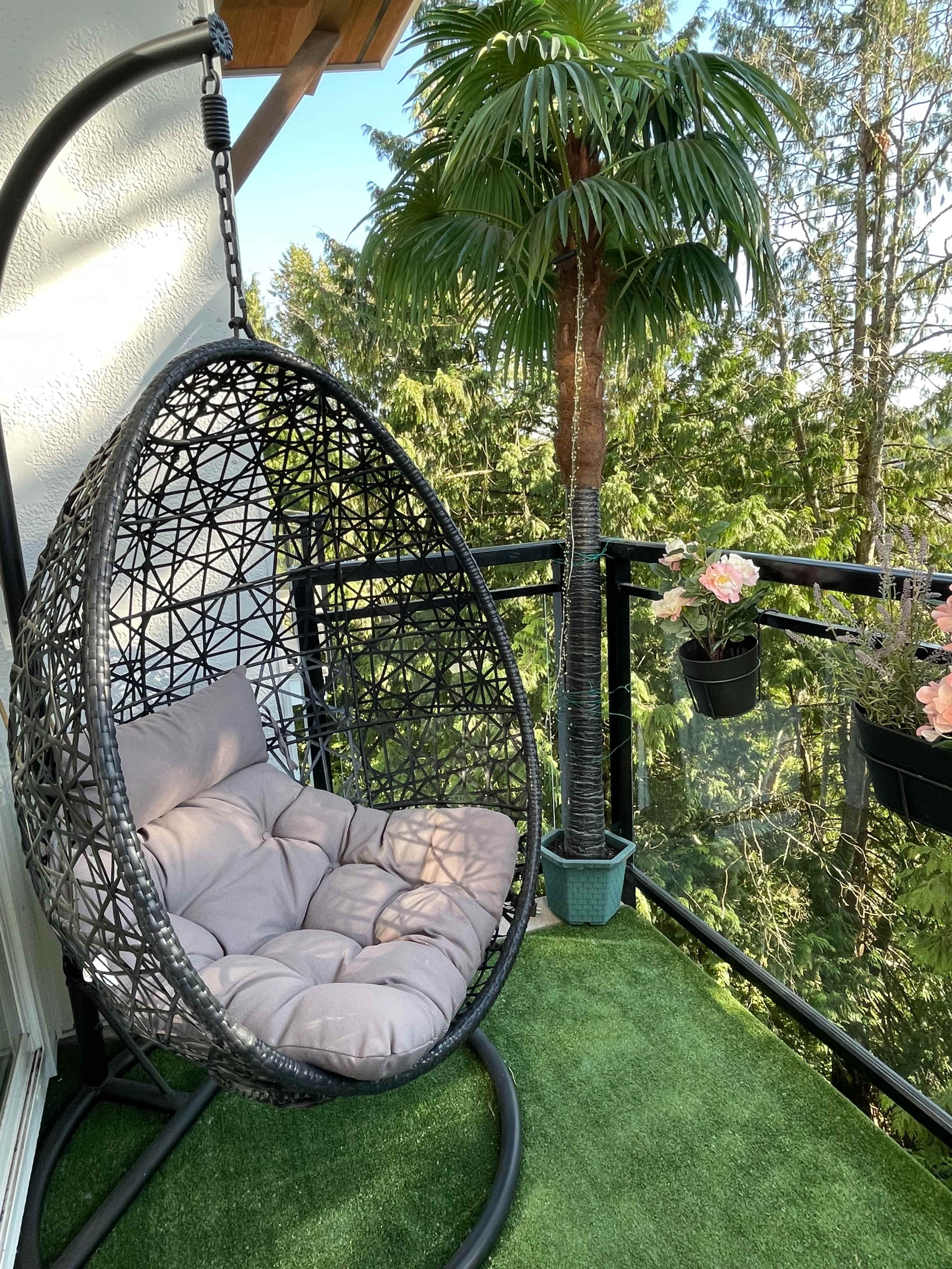 A hanging chair with cushions is positioned on a balcony surrounded by greenery and potted plants.