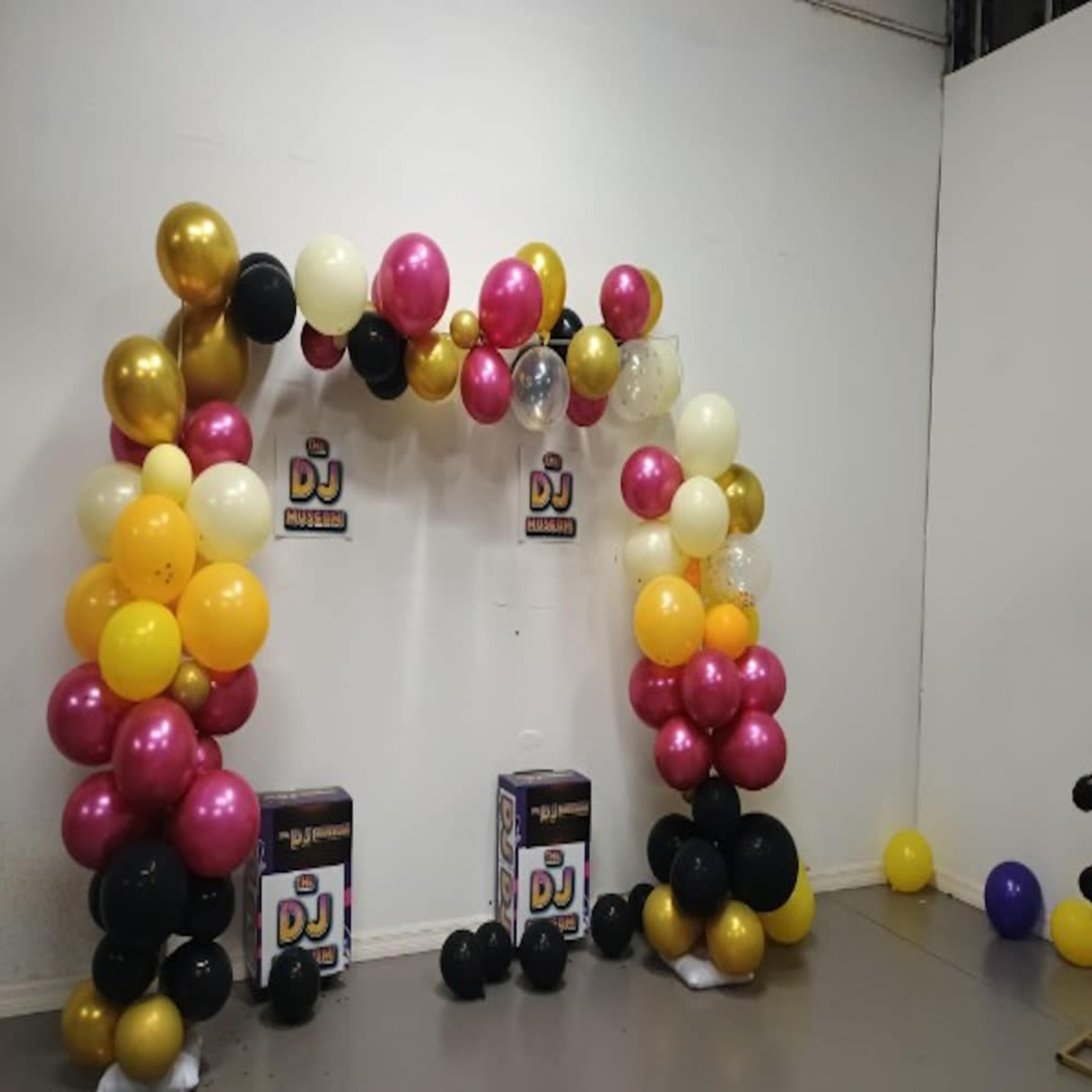 The image shows a balloon arch decorated with pink, gold, black, and white balloons, with DJ-themed signage at the center.