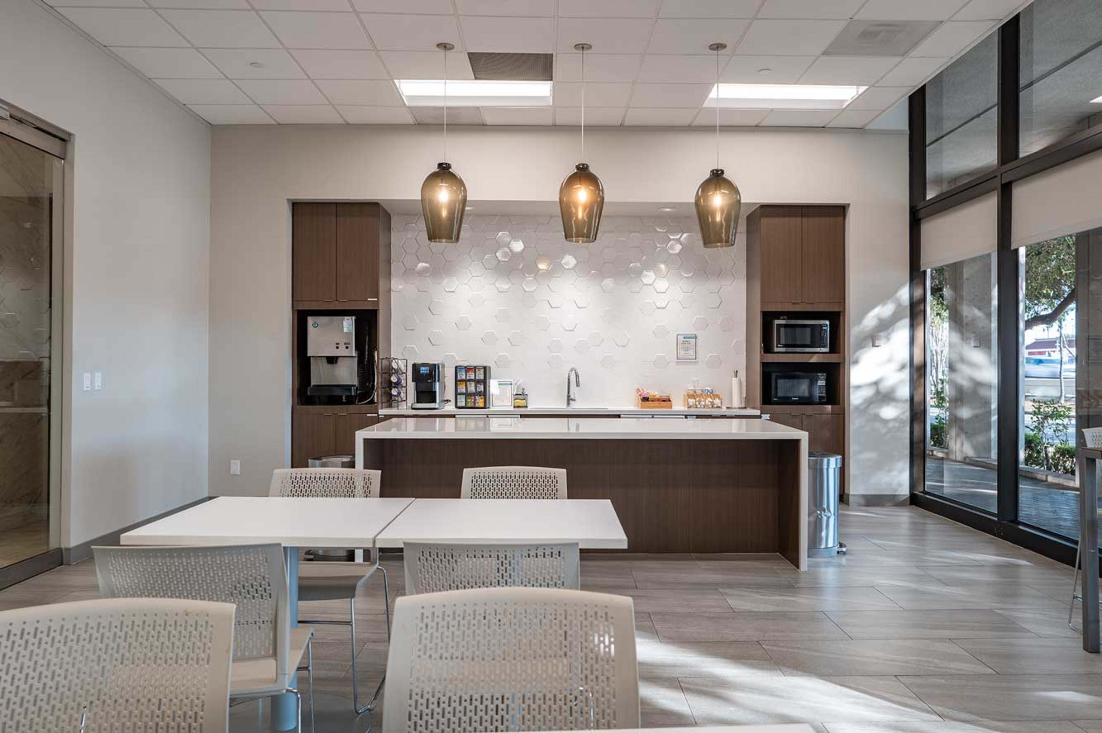 The image shows a modern kitchenette area with a counter, coffee machine, and appliances, accompanied by several tables and chairs.