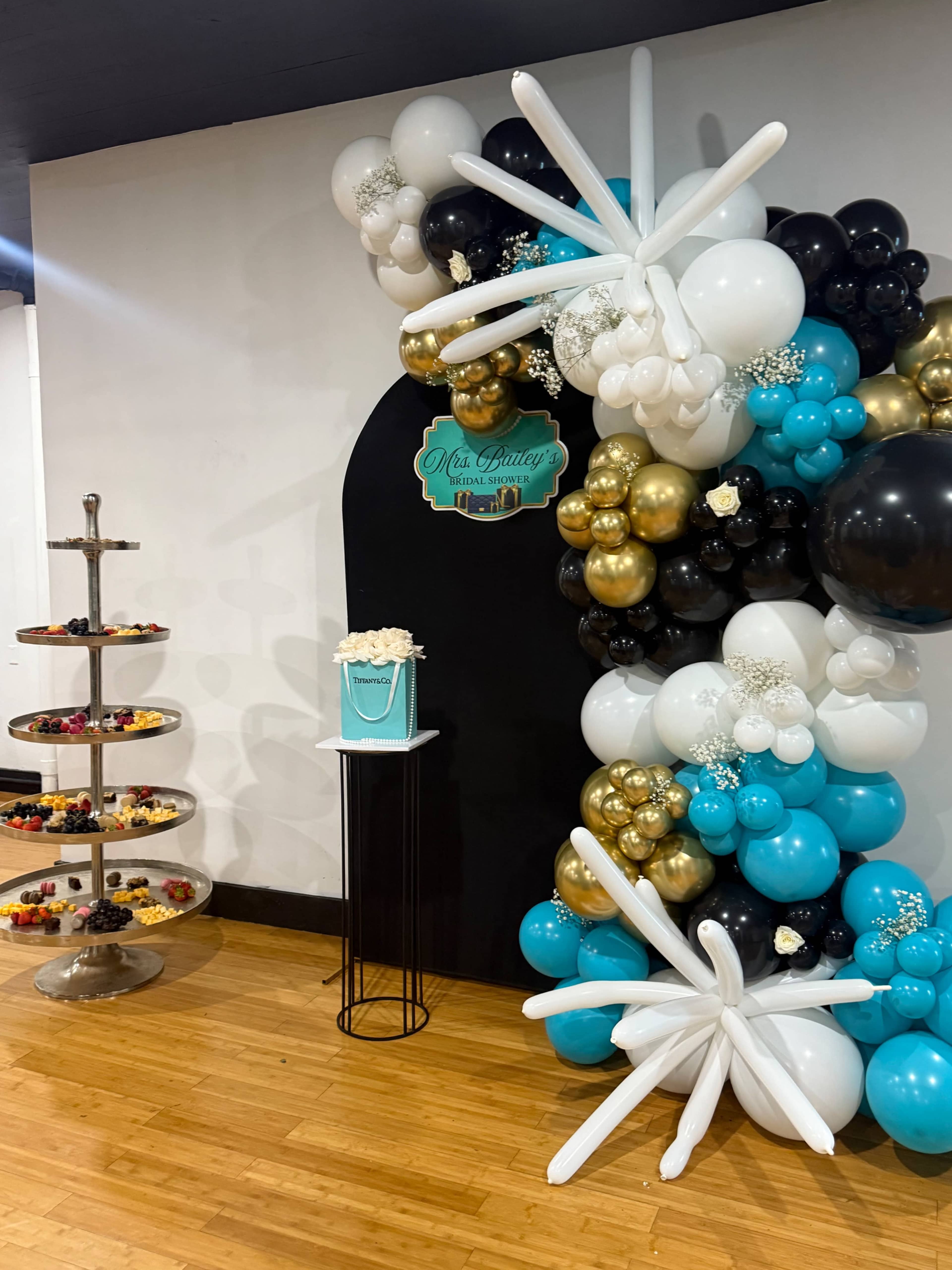 A decorative display features an arch of balloons in various colors alongside a tiered stand filled with assorted pastries.