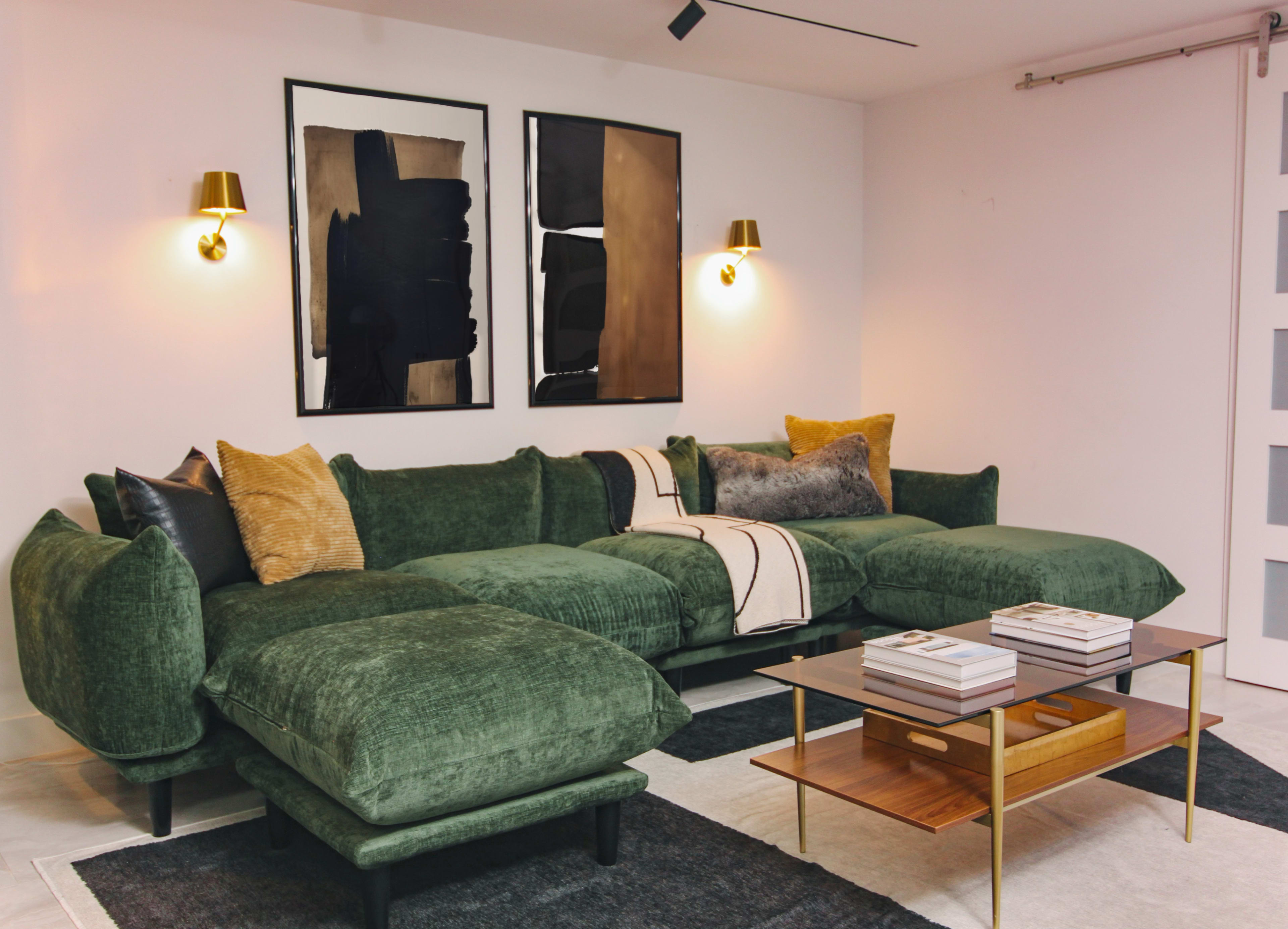 A modern living room features a large green sectional sofa, two abstract framed artworks on the wall, and a wooden coffee table on a dark area rug.