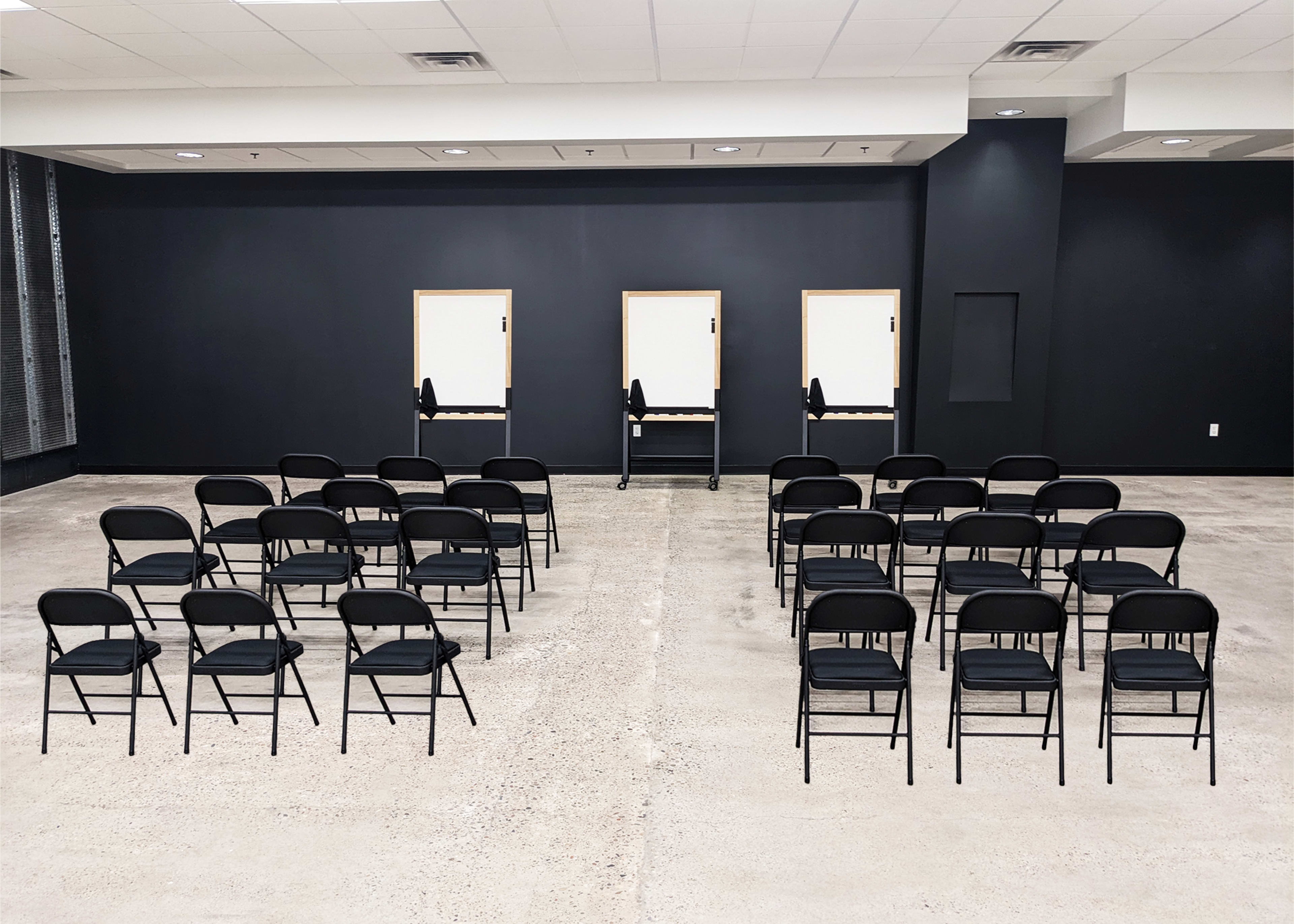 A spacious room with black walls, arranged with rows of black folding chairs facing three blank podiums.