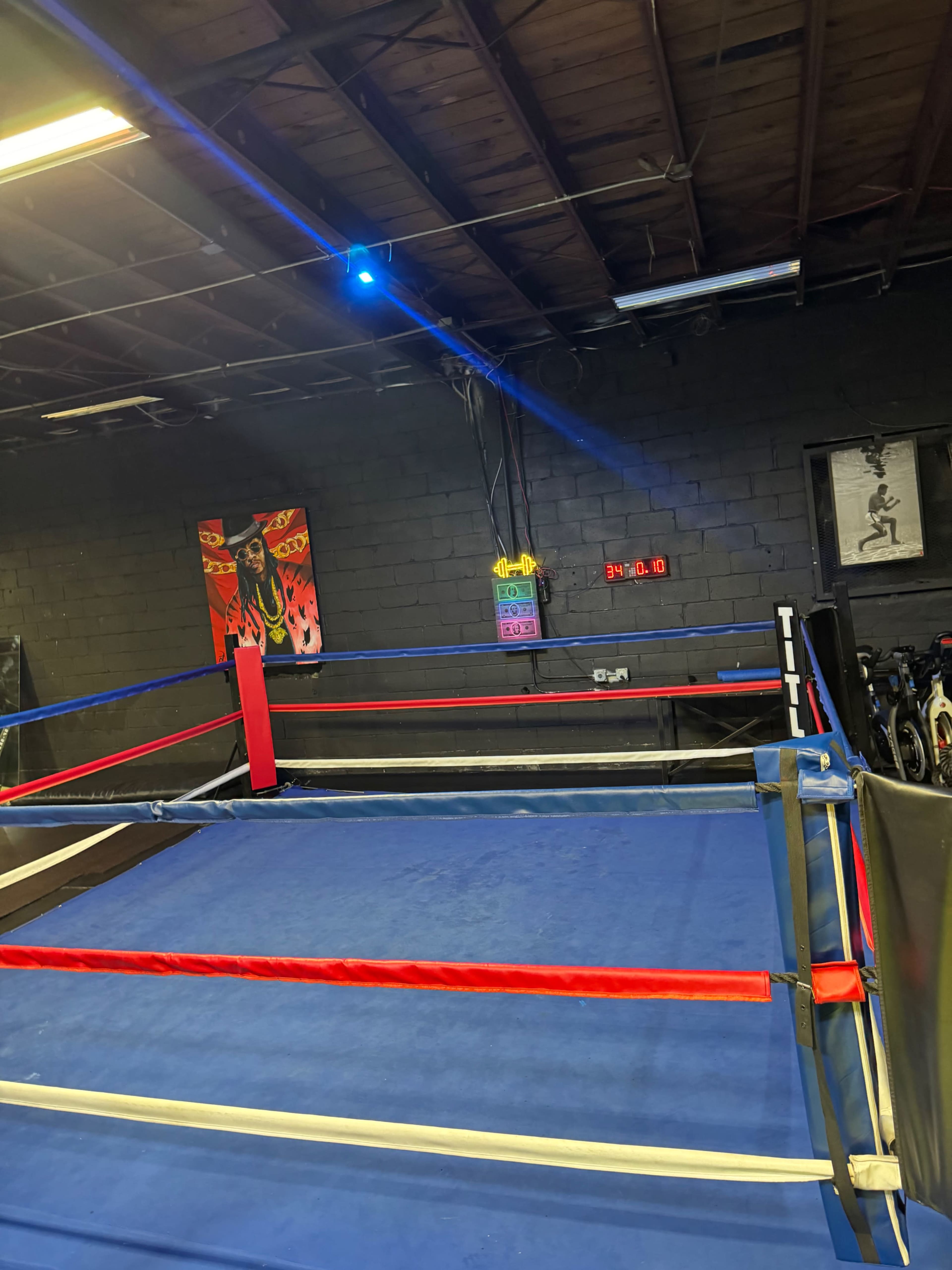 The image shows an empty boxing ring with blue mats, red and blue corner posts, and gym equipment in the background.