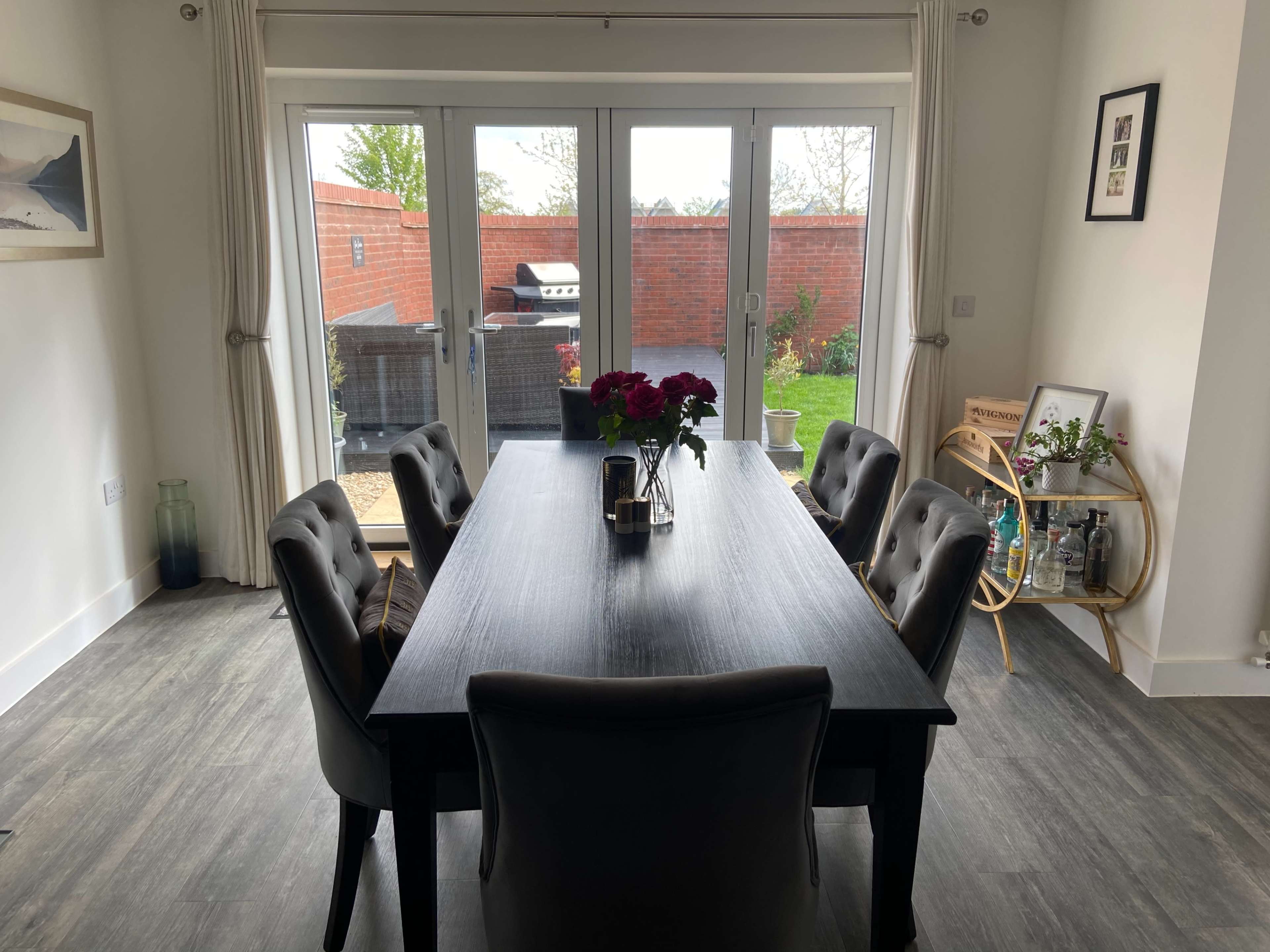 A dining table with dark chairs is set in a room overlooking a garden through large glass doors.