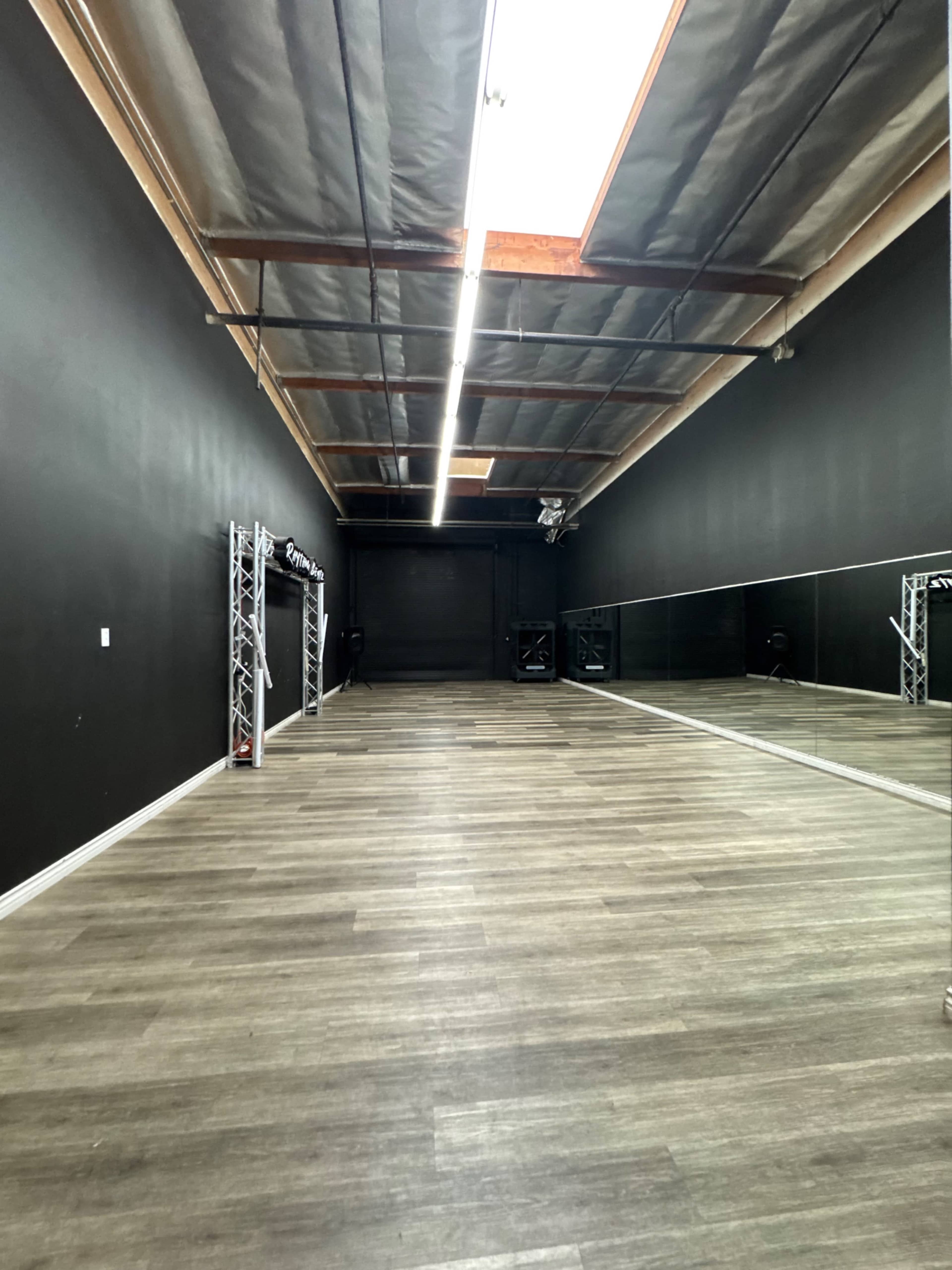 The image shows a spacious dance studio with a wooden floor, black walls, and a long mirror lining one side.