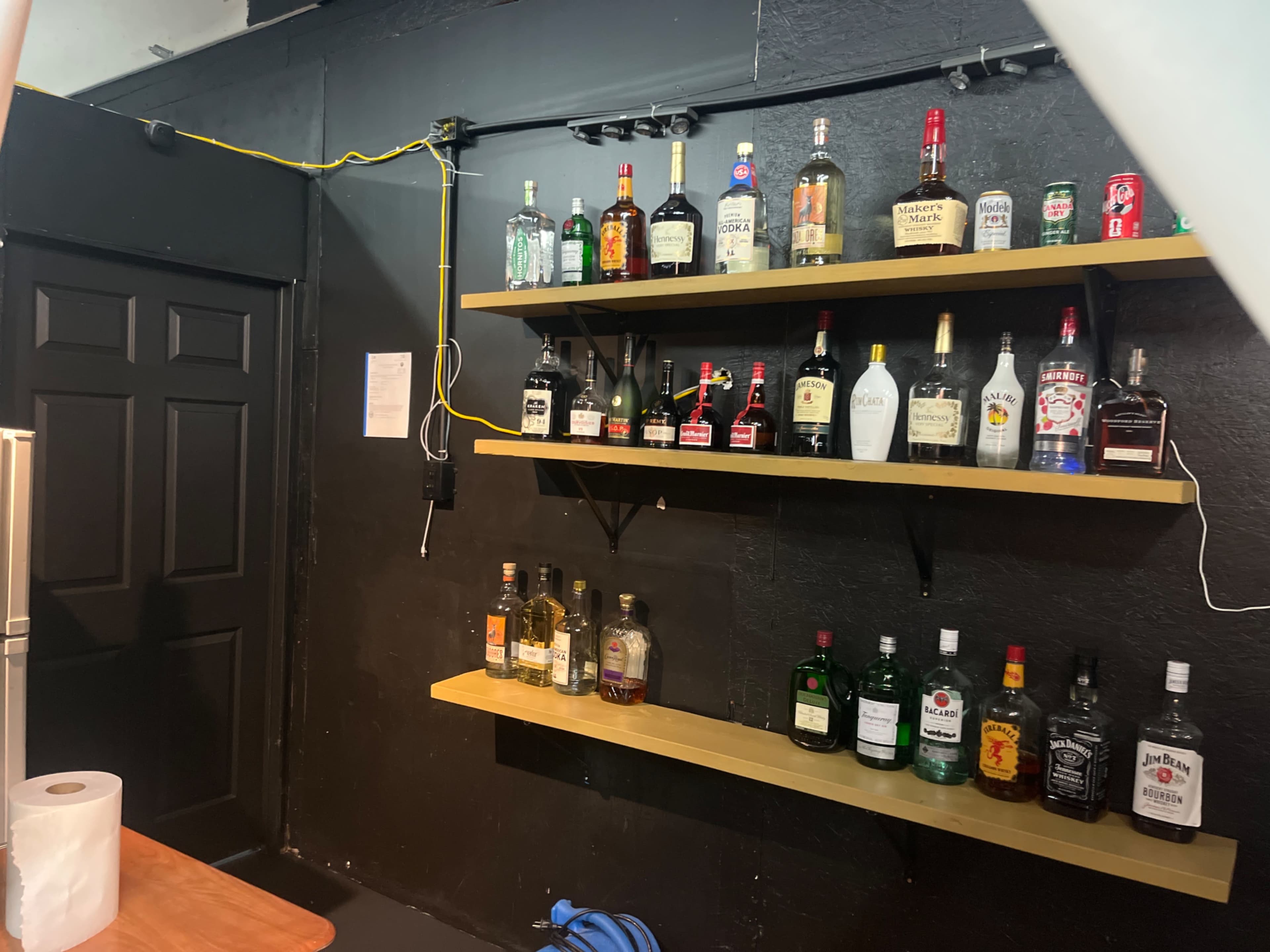 The image shows a black wall with two wooden shelves displaying various bottles of liquor, including spirits and liqueurs.