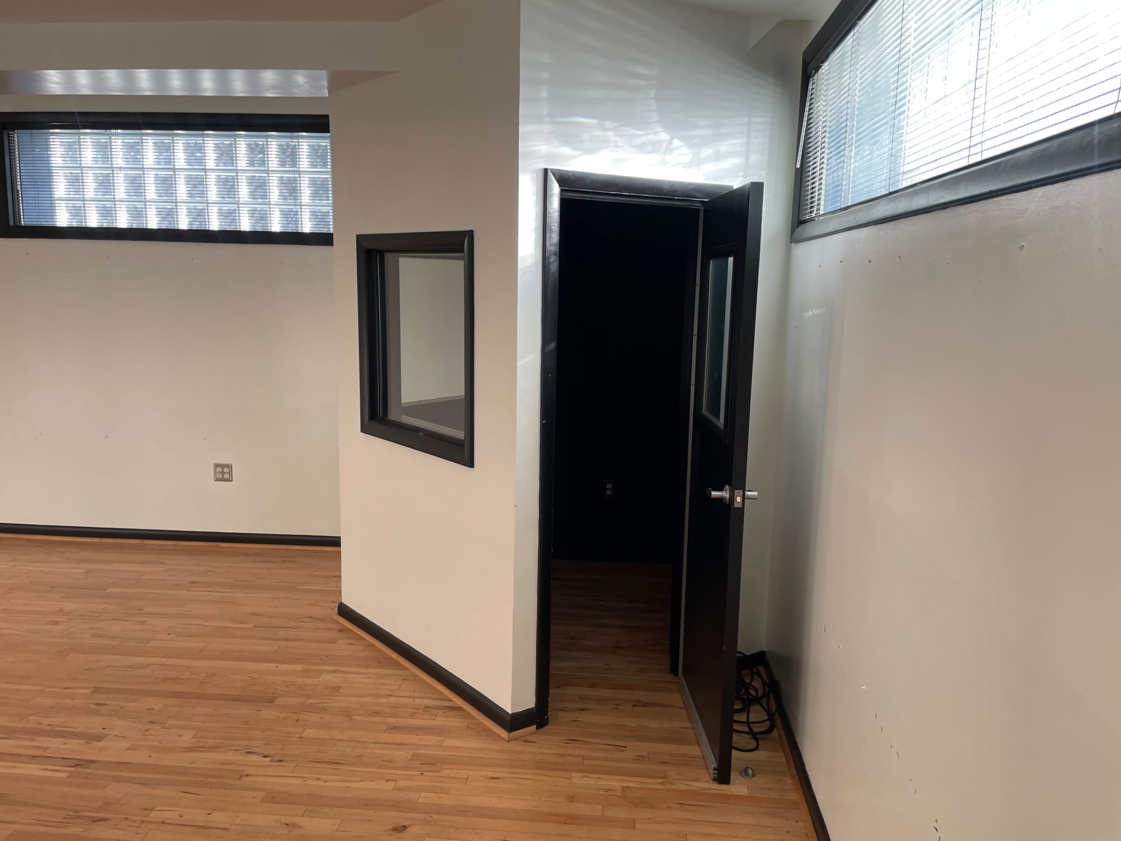 The image shows a room with wooden flooring, featuring a door on the right that opens to a darkened space and a window on the left.