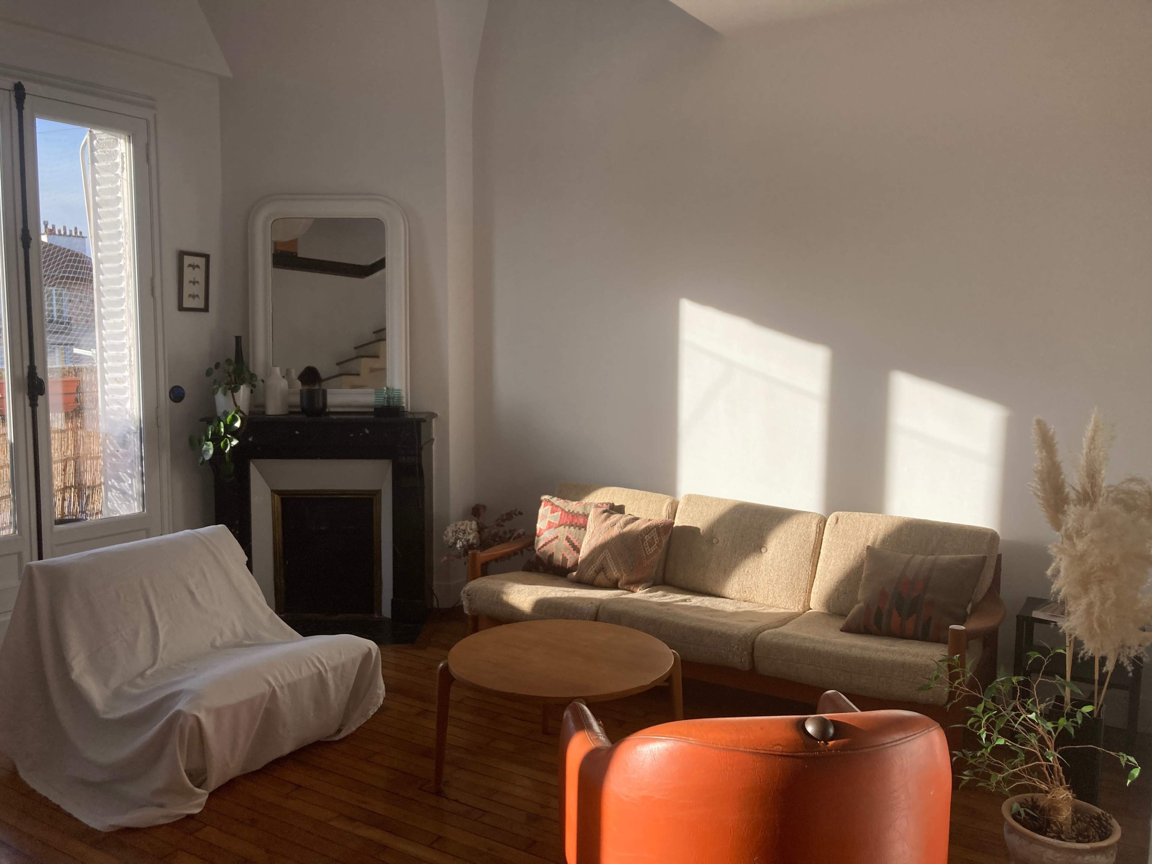 The living room features a sofa, a round coffee table, and a chair, with sunlight streaming through a window and casting shadows on the walls.