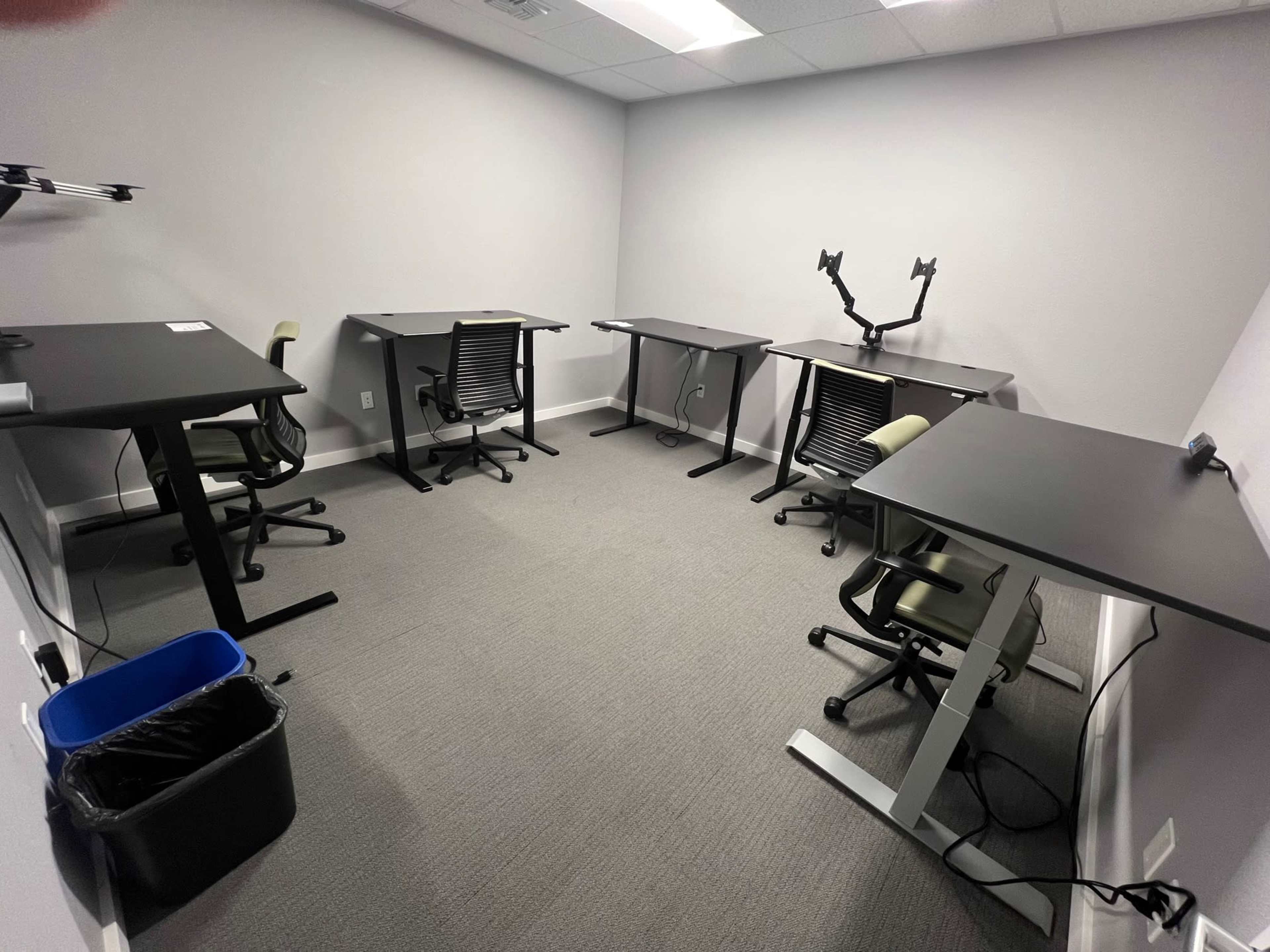 A small office space with five adjustable desks and chairs arranged around the room, along with a trash bin in the corner.
