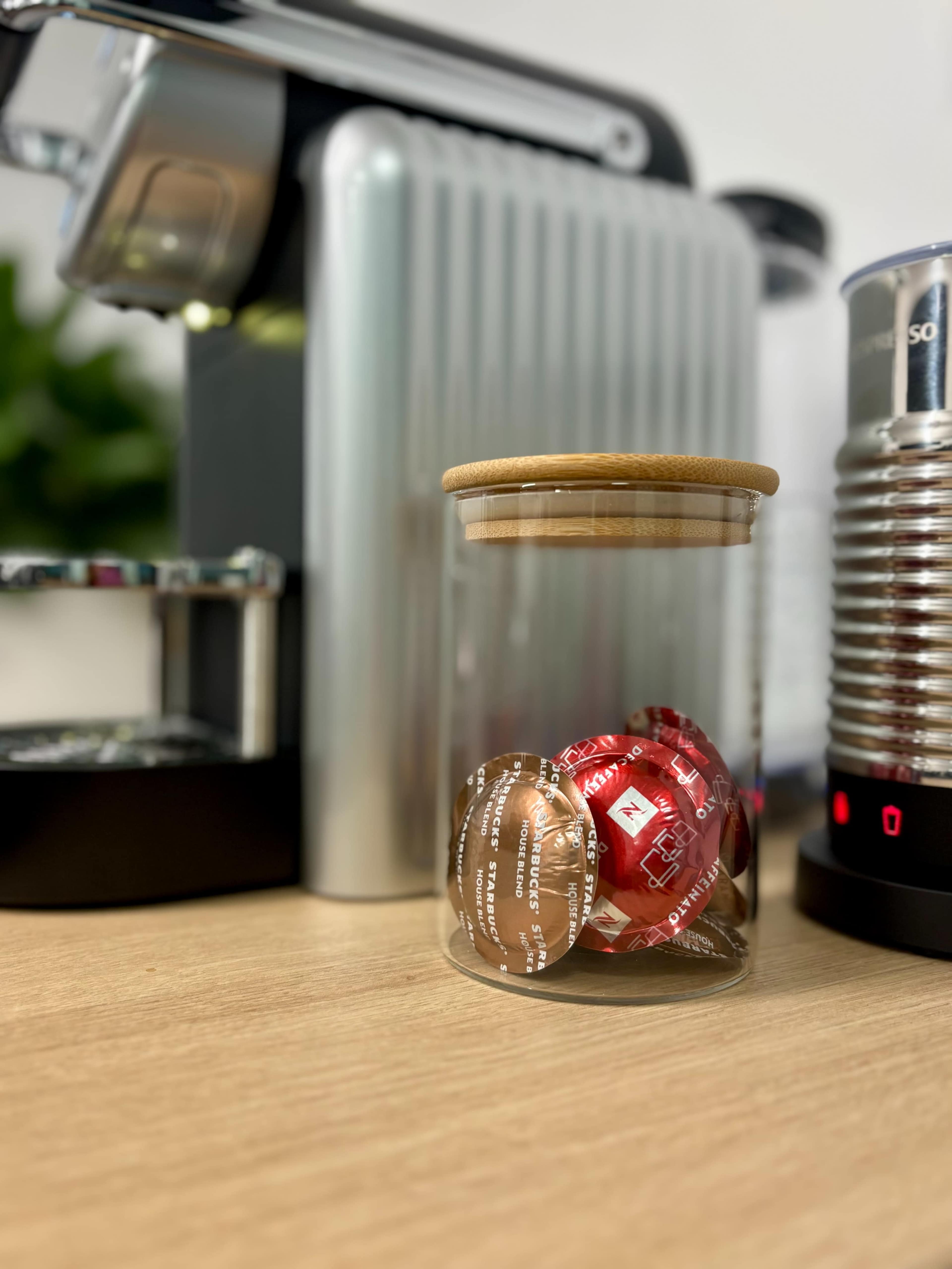 A glass jar with a bamboo lid contains coffee capsules, placed on a wooden table near an espresso machine and a frother.