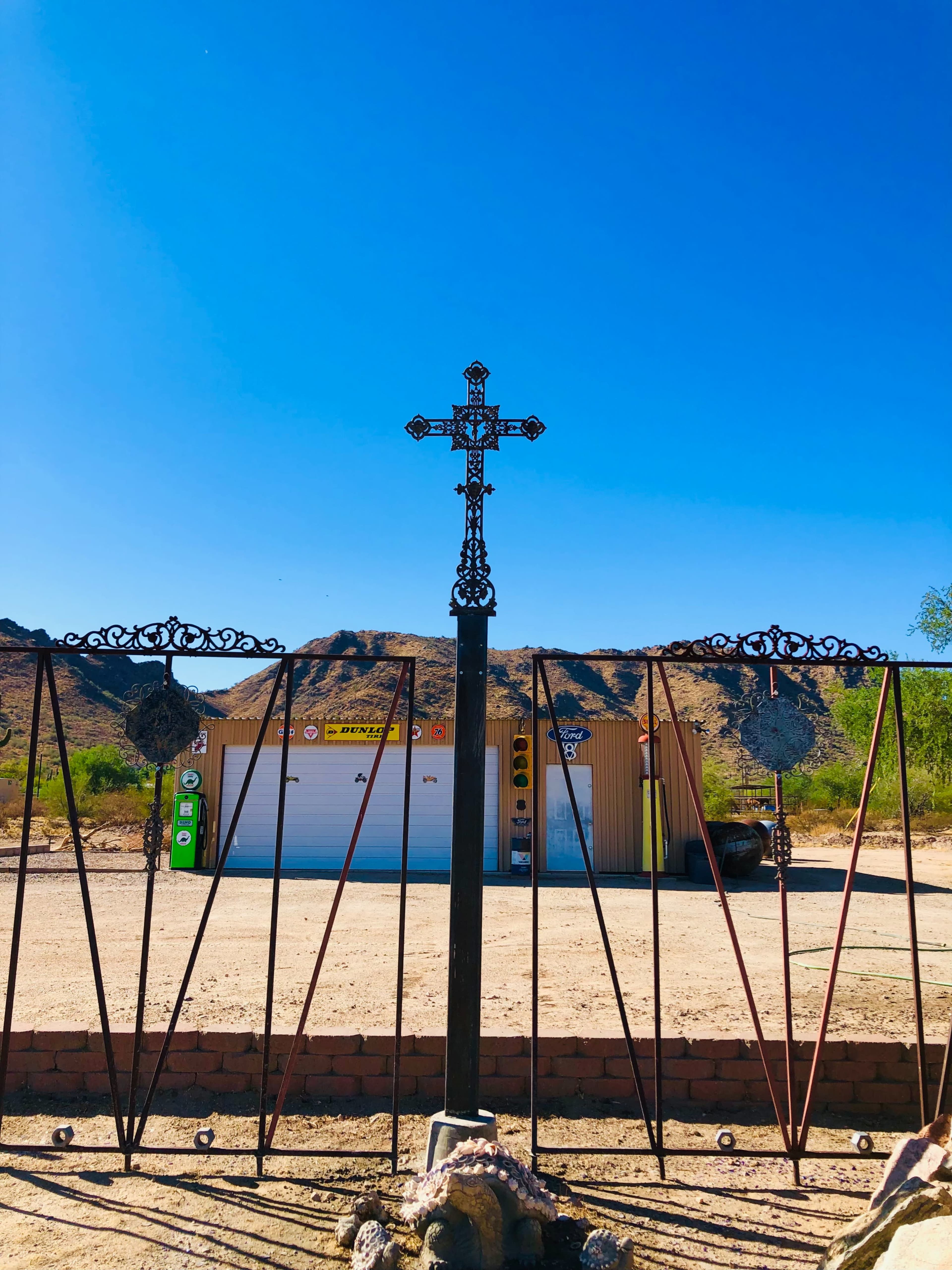 A decorative iron cross stands at the center of a gated entrance, with a building and mountains visible in the background under a clear blue sky.